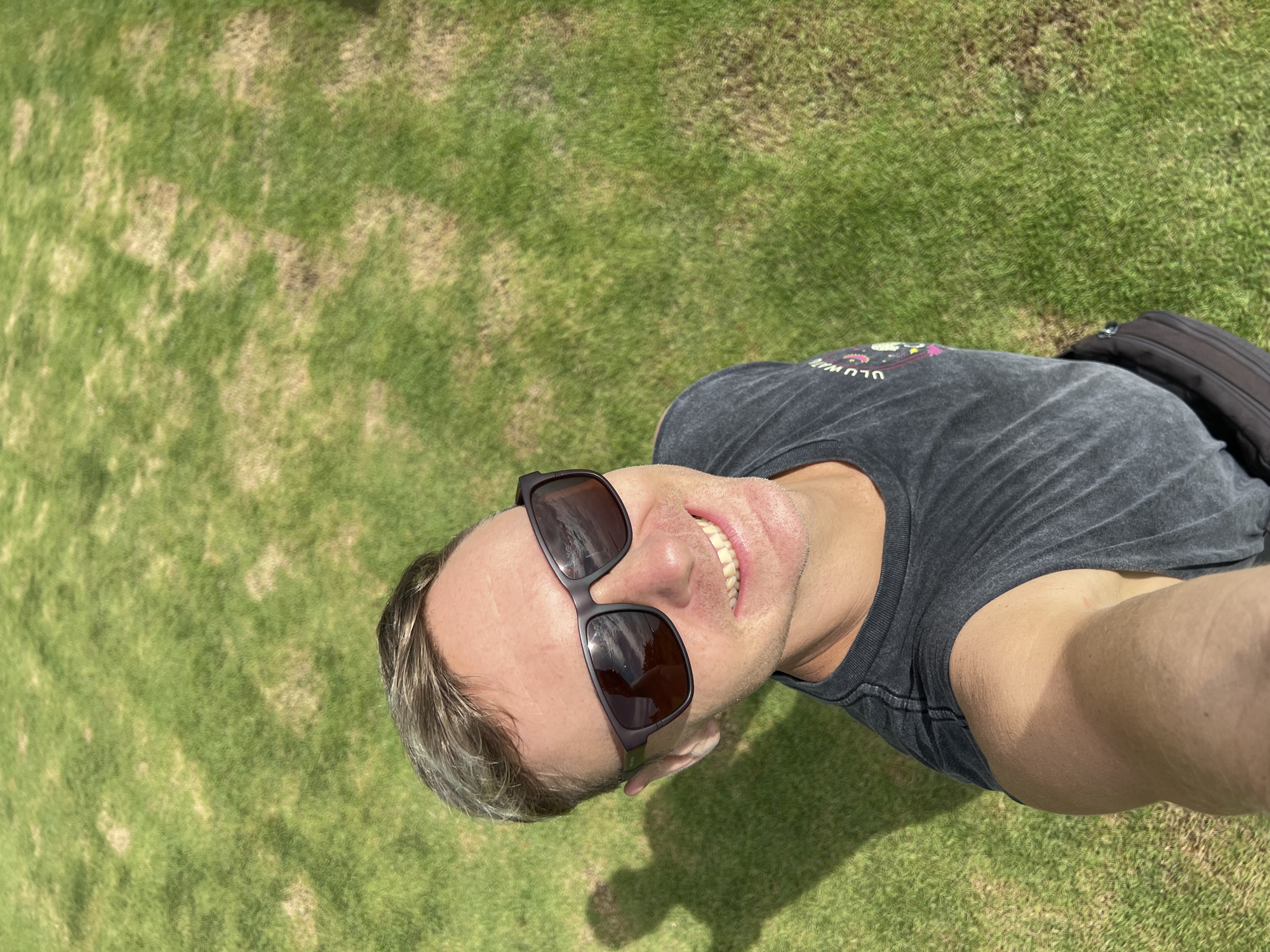 A man wearing sunglasses and a gray t-shirt is standing on a grassy area. The grass appears to be somewhat dry in places, suggesting a sunny environment. The man is holding the camera at an angle, capturing a selfie-style shot. The presence of grass could indicate a park or golf course location in Kuta Selatan, Indonesia.