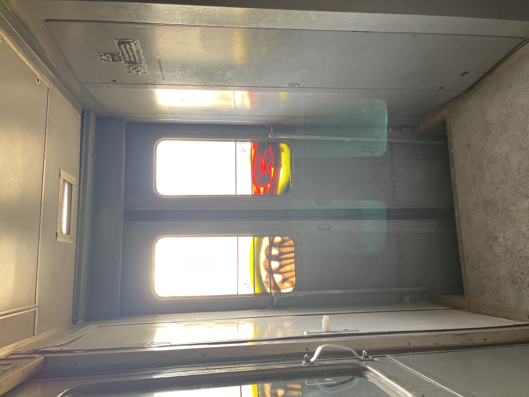 The image shows the interior of a subway car in Kyiv, Ukraine, featuring two sets of double doors leading to the outside. The doors have windows with colorful, stained-glass-like inserts depicting abstract patterns. Sunlight streams through the windows, creating bright flares across the scene. The walls and doors are painted a muted, industrial gray. A metal handle is visible on the left side, and a small warning sign is affixed to the right wall. The floor is concrete and appears worn. There is an opening in the floor between the doors, suggesting a step down. No people are visible in the image.