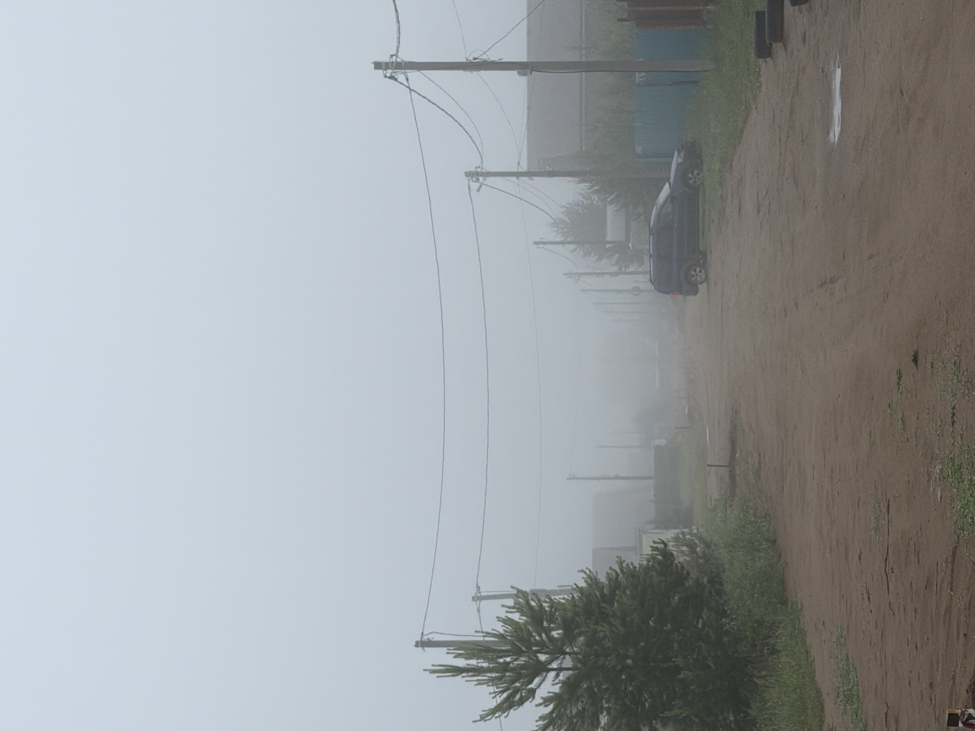The image depicts a quiet, heavily fogged scene of a dirt road extending into the distance in what appears to be a rural or semi-urban setting in Chaikovsky, Russia. The dense white fog significantly reduces visibility, enveloping the landscape in a muted, hazy atmosphere.
Along the left side of the unpaved road, a series of wooden utility poles with overhead power lines recede into the thick mist, becoming progressively fainter. A dense, dark green evergreen bush with needle-like leaves occupies the lower left foreground.
On the right side of the dirt road, a dark-colored car, possibly a sedan or a small SUV, is parked facing away from the viewer. Its rear is visible, but the rest of the vehicle is largely obscured by the fog. Further down the road on the right, several indistinct dark cylindrical objects, possibly tires or bins, are faintly visible. The road itself is composed of packed dirt, with patches of green grass and other sparse vegetation growing along its edges and shoulders. Faint outlines of structures or buildings can be seen in the far distance, heavily blurred by the pervasive fog.
The heavy fog is the most prominent feature, creating a sense of stillness and isolation. The time of day is difficult to ascertain precisely due to the lack of clear sunlight, but it appears to be daytime. There are no people visible, and no specific activity is taking place beyond the stationary car. No legible text is present in the image.