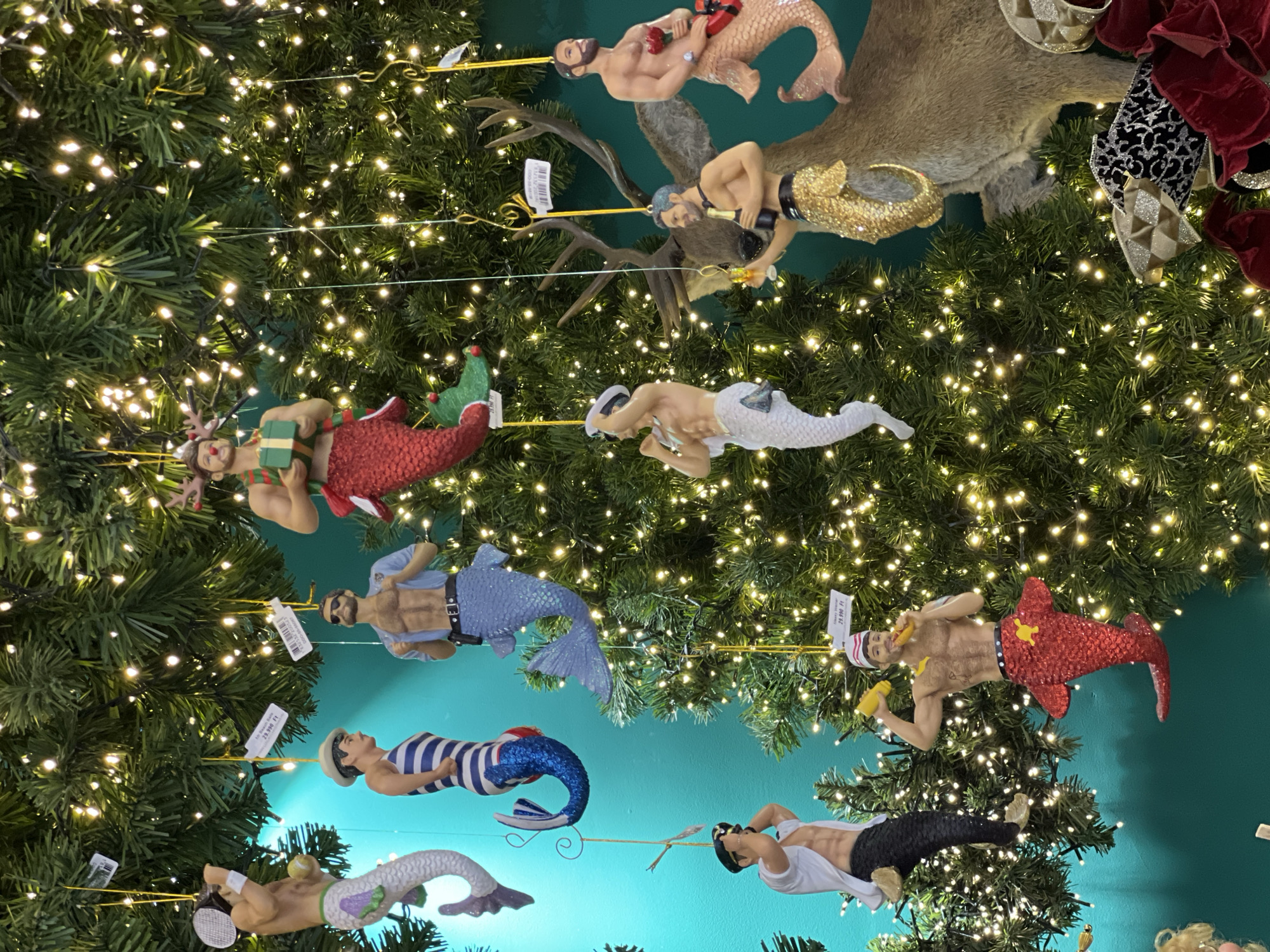 The image shows a decorated Christmas tree with a collection of male mermaid ornaments. The mermen have various skin tones and tail colors, including red, blue, white, and black. They are dressed in a variety of outfits, including a sailor costume, a police uniform, and tennis attire. The tree is heavily adorned with lights and has a taxidermied deer head mounted near the top. There is a display of ornaments and decorations visible on the upper right side of the tree. The background wall appears to be teal-colored.
The photograph may have been taken in Budapest, Hungary as a possibility but this is not certain.