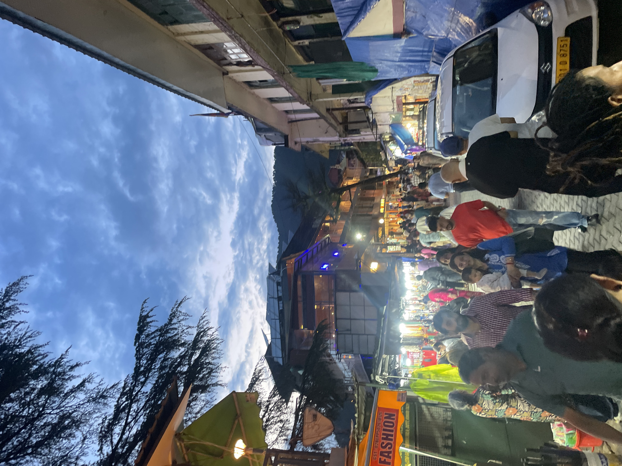 The image depicts a bustling street scene in Dharamshala, India, during twilight. The sky above is a deep blue with scattered clouds, suggesting the transition from day to night. The street is lined with buildings, some appearing to be shops with illuminated signs, like one prominently displaying "FASHION" in red lettering on an orange background. Other shops appear to have goods displayed under awnings or inside. A diverse crowd of people, dressed in casual attire, are walking along the street, suggesting a lively marketplace or thoroughfare. Among the people, a woman is holding a child, and a man in a red shirt is visible. Several vehicles, including a white car with a yellow license plate, are present on the street, indicating it's a functional urban area. The presence of trees with dark foliage framing the upper left corner adds a natural element to the urban environment. The overall atmosphere suggests a vibrant evening in a commercial district.
