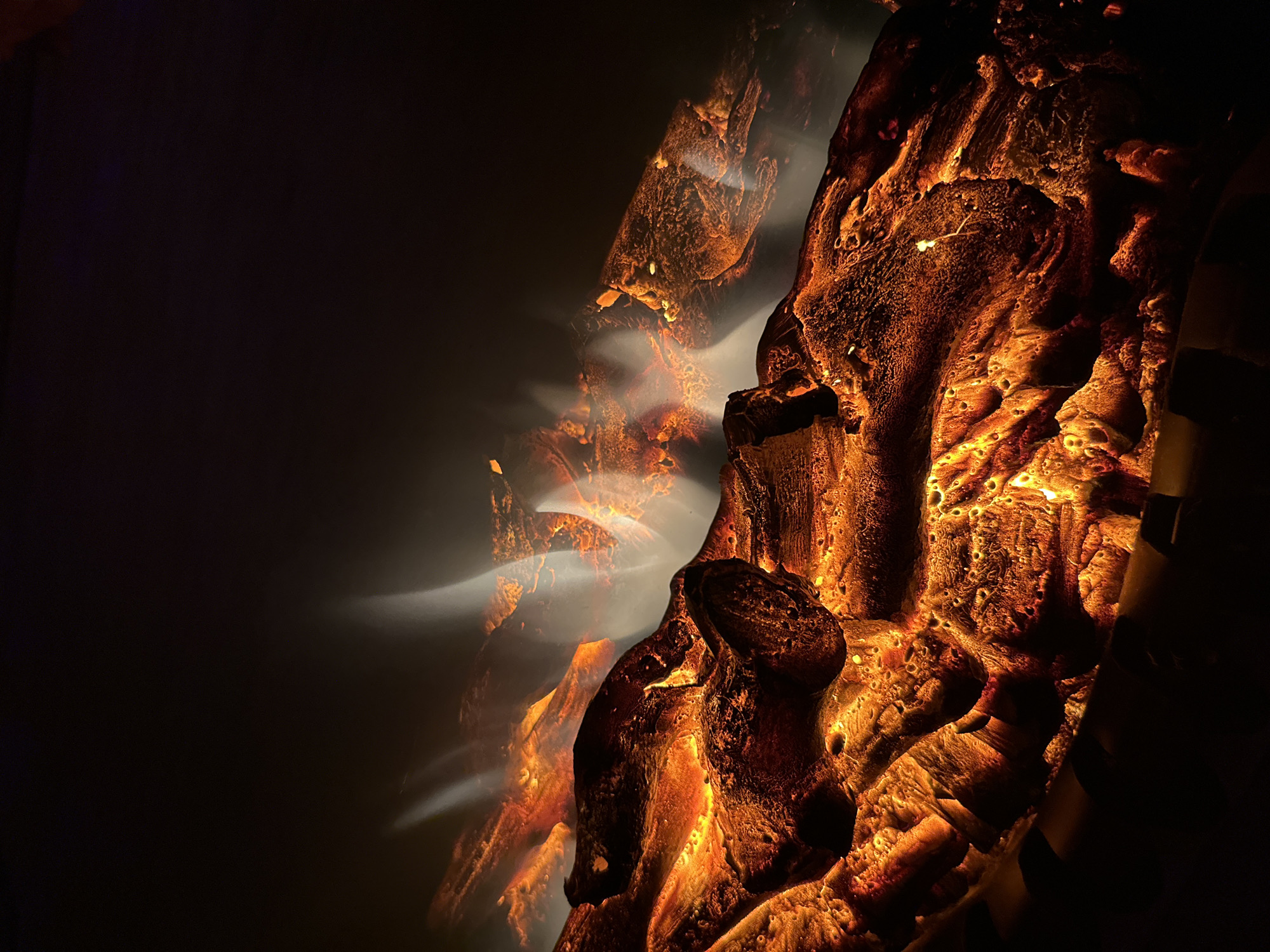 The image shows a close-up, low-angle shot of a fire pit or fireplace. The primary focus is on logs that appear to be glowing with an intense orange and yellow light, simulating embers. Wisps of white and grey smoke or light are rising from the logs, adding to the illusion of a fire. The background is dark, creating a stark contrast and enhancing the luminescence of the logs. The texture of the logs is rough and detailed, showing crevices and uneven surfaces. Some parts of the logs are brighter than others, suggesting varying heat intensity. The overall impression is one of warmth and simulated combustion within a dark environment.