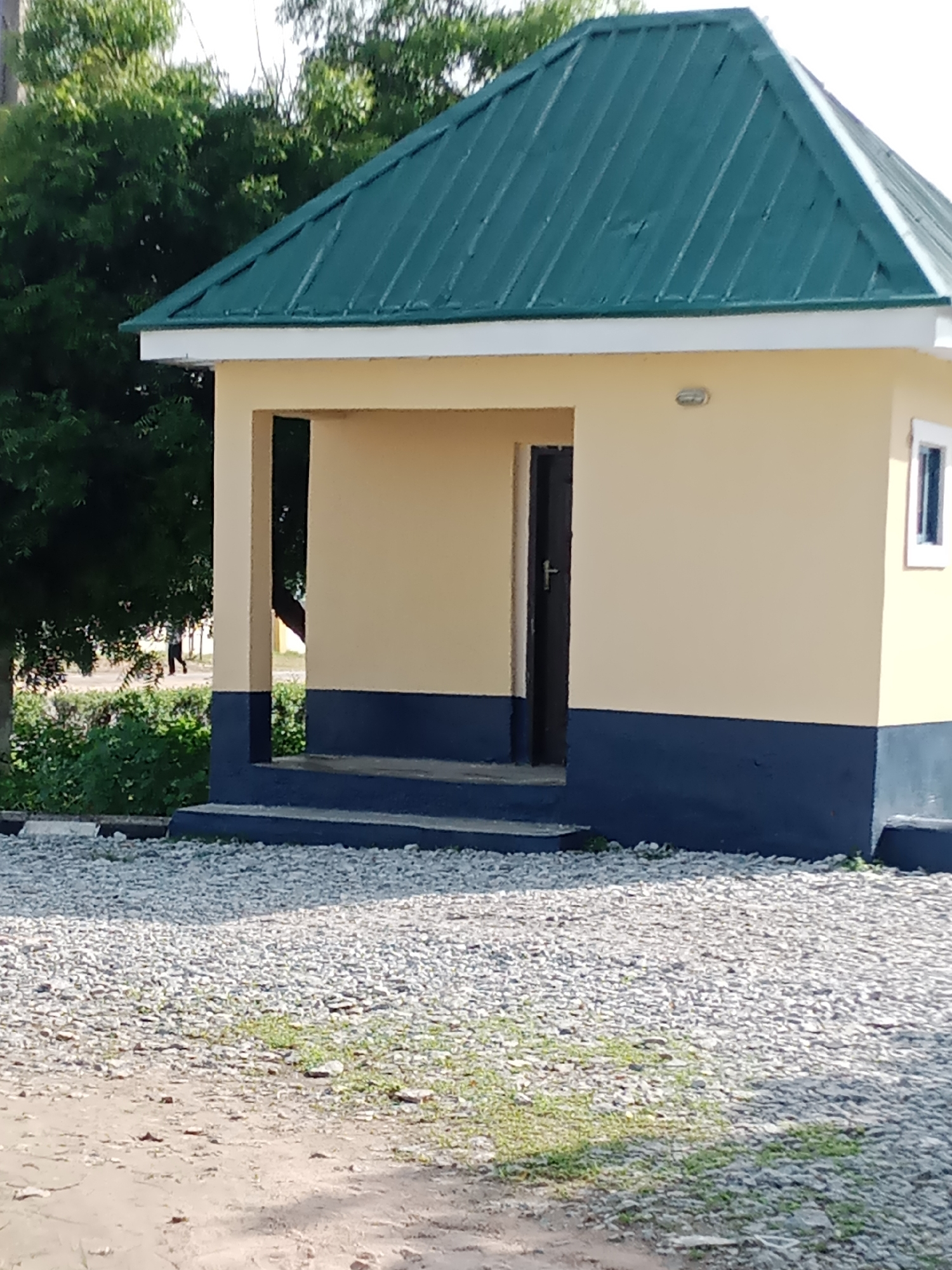 The image shows a small, modern building with a peaked green metal roof and pale yellow walls. The lower portion of the building is painted dark blue. There is an open doorway to the left, leading into darkness, with a dark brown door visible inside. A single small window with a white frame is visible on the right side of the building. The building is set on a gravel and dirt surface, with some green vegetation visible in the background and to the left. In the distance, a person is walking away from the camera towards the left. The overall impression is of a functional, possibly public, building in an outdoor urban or semi-urban setting. The location is Jalingo, Nigeria.