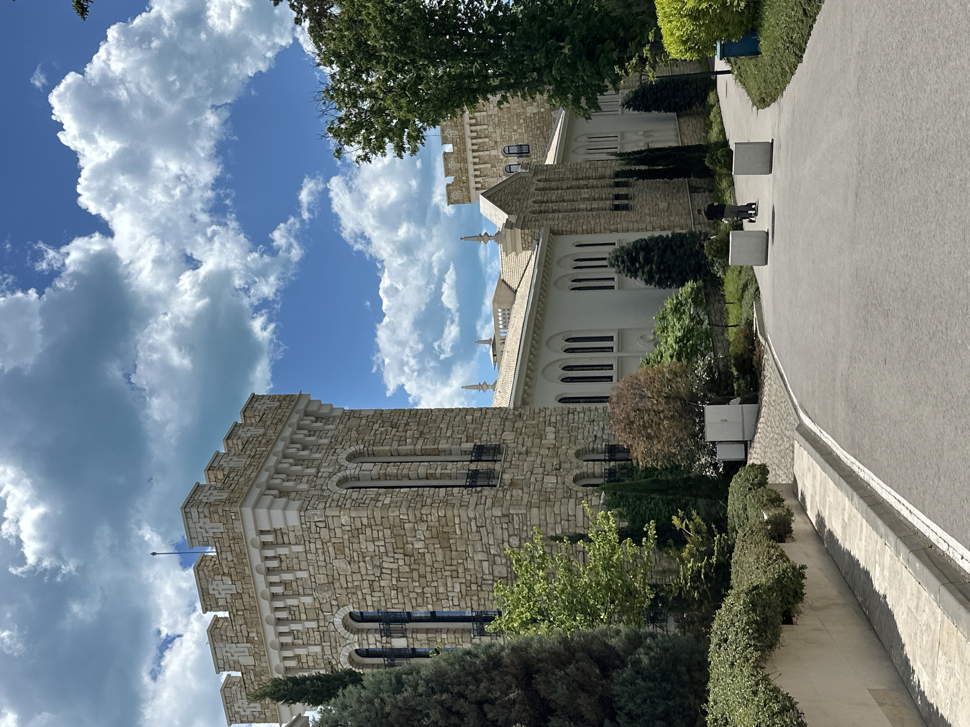 The image shows a building complex with stone and white concrete architectural elements, likely a government or cultural institution, set against a blue sky with puffy white clouds. The building features a tower with an uneven top and arched windows, as well as other structures with white facades and rows of windows. A paved walkway runs alongside the building, lined with trimmed bushes and square concrete planters. A person with a stroller can be seen on the walkway. The location is likely in Varna, Bulgaria.