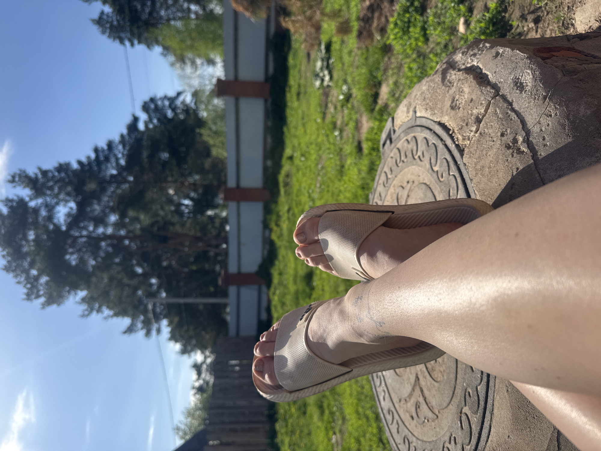 The image shows a pair of bare legs wearing beige sandals, resting on a circular concrete structure with a decorative pattern. The background includes a wooden fence, green grass, trees, and a blue sky. The location is purportedly Rusalevka, Russia. No specific actions or interactions are depicted.