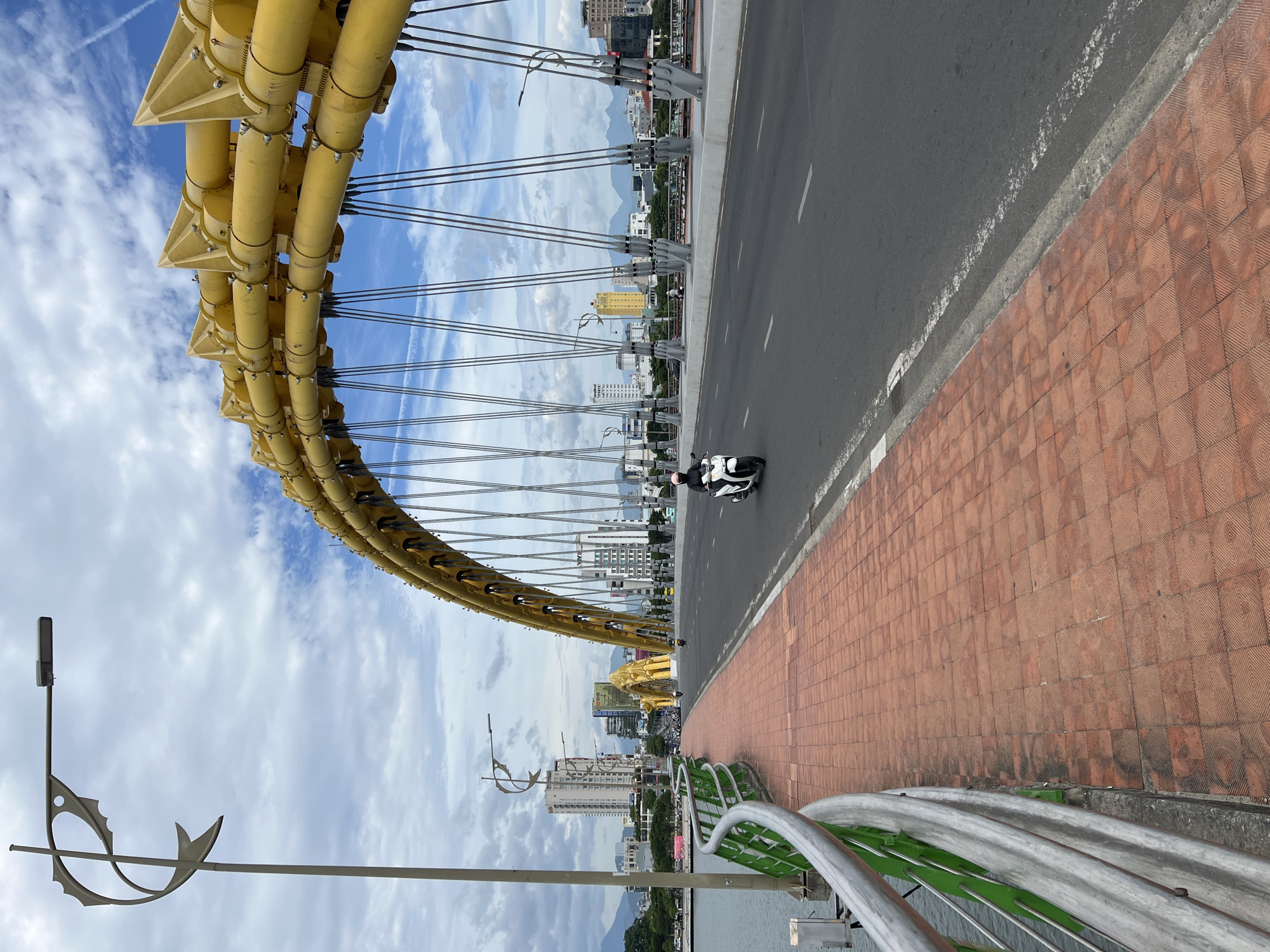 This image depicts the Dragon Bridge (Cầu Rồng) in Đà Nẵng, Vietnam, spanning the Han River. The perspective is from the pedestrian sidewalk on the left side of the bridge, looking towards the city's east bank.
In the immediate foreground on the left, a metal guardrail with silver top rails and green vertical bars separates the sidewalk from the river, the surface of which is visible in the bottom left corner. Adjacent to the guardrail is a pedestrian walkway paved with red-orange square tiles featuring a concentric square pattern. A tall, light-colored streetlamp with a distinctive stylized fish-shaped ornamentation is positioned on the far left.
The midground features an asphalt roadway extending from the bottom right towards the background. A white broken line separates the main traffic lane from the curb alongside the pedestrian path. A white scooter with a single rider wearing a dark helmet is visible on the roadway, moving away from the viewer. The main structure of the Dragon Bridge dominates the upper midground and background, characterized by its prominent yellow segmented arch and numerous gray steel suspension cables that connect the arch to the bridge deck. The segments of the arch appear to be bolted together, forming a dragon-like spine.
In the background, the urban landscape of Đà Nẵng unfolds, with various multi-story buildings of differing heights and architectural styles, predominantly in white, grey, and some with reflective glass facades. A notable yellow high-rise building is visible further down the bridge. Distant mountains are discernible on the horizon beneath a partly cloudy sky, showing patches of blue interspersed with white and light grey clouds. Faint contrails are present in the sky above the bridge's arch.