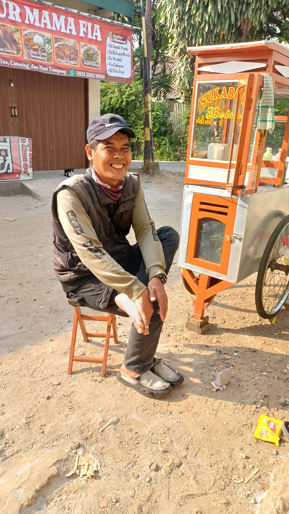 A man sits on a small wooden stool in front of a food cart in Gunung Putri, Indonesia. He is wearing a black baseball cap, a light brown long-sleeved shirt with dark gray vest, dark pants, and gray slip-on shoes. He is smiling and looking towards the camera. The food cart is made of orange and silver metal, with a sign that says "SUKABUMI BERKAH" in orange letters. There is a wheel on the right side of the cart. In the background, there is a building with a brown door and a banner that says "UR MAMA FIA". There are also trees and a sunny sky. The ground is covered with dirt and small rocks. A small black cat is visible near the brown door.