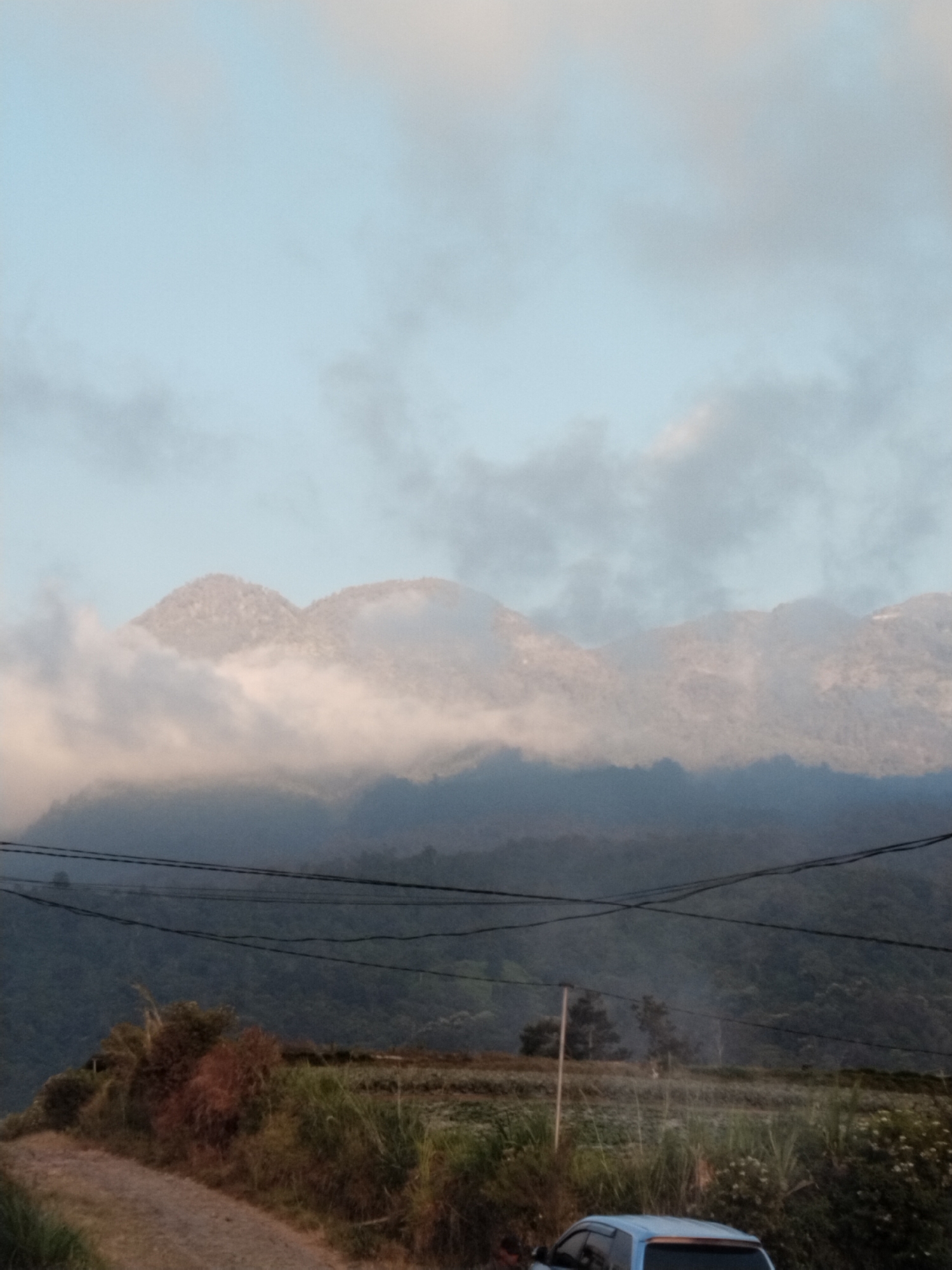 The image depicts a scenic view of mountains shrouded in mist, possibly taken in the area of Gumeng, Indonesia. The foreground features a dirt path leading into the frame, bordered by dry grass and a few bushes. Power lines crisscross the scene horizontally in the middle ground, separating the foreground from the lush green mountains in the background. A portion of a light blue vehicle is visible in the lower right corner, suggesting it is parked near the path. The sky above is a pale blue, with scattered clouds adding depth to the atmospheric perspective.