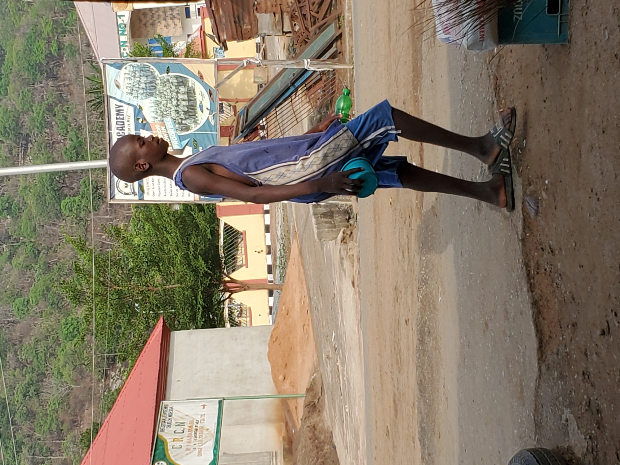 A young, bald, dark-skinned boy walks barefoot along a cracked pavement in Mararaba, Nigeria. He carries a blue bowl in one hand and a green bottle in the other. He wears a sleeveless blue shirt with a white stripe and matching blue shorts. Behind him, an advertising billboard for an academy can be seen next to some lush green trees and an elevated rocky terrain with a mountain in the background. Adjacent to the boy, a sign for the Christian Reformed Church of Nigeria (CRCN) can be seen. The pavement is rough and shows signs of wear and tear.