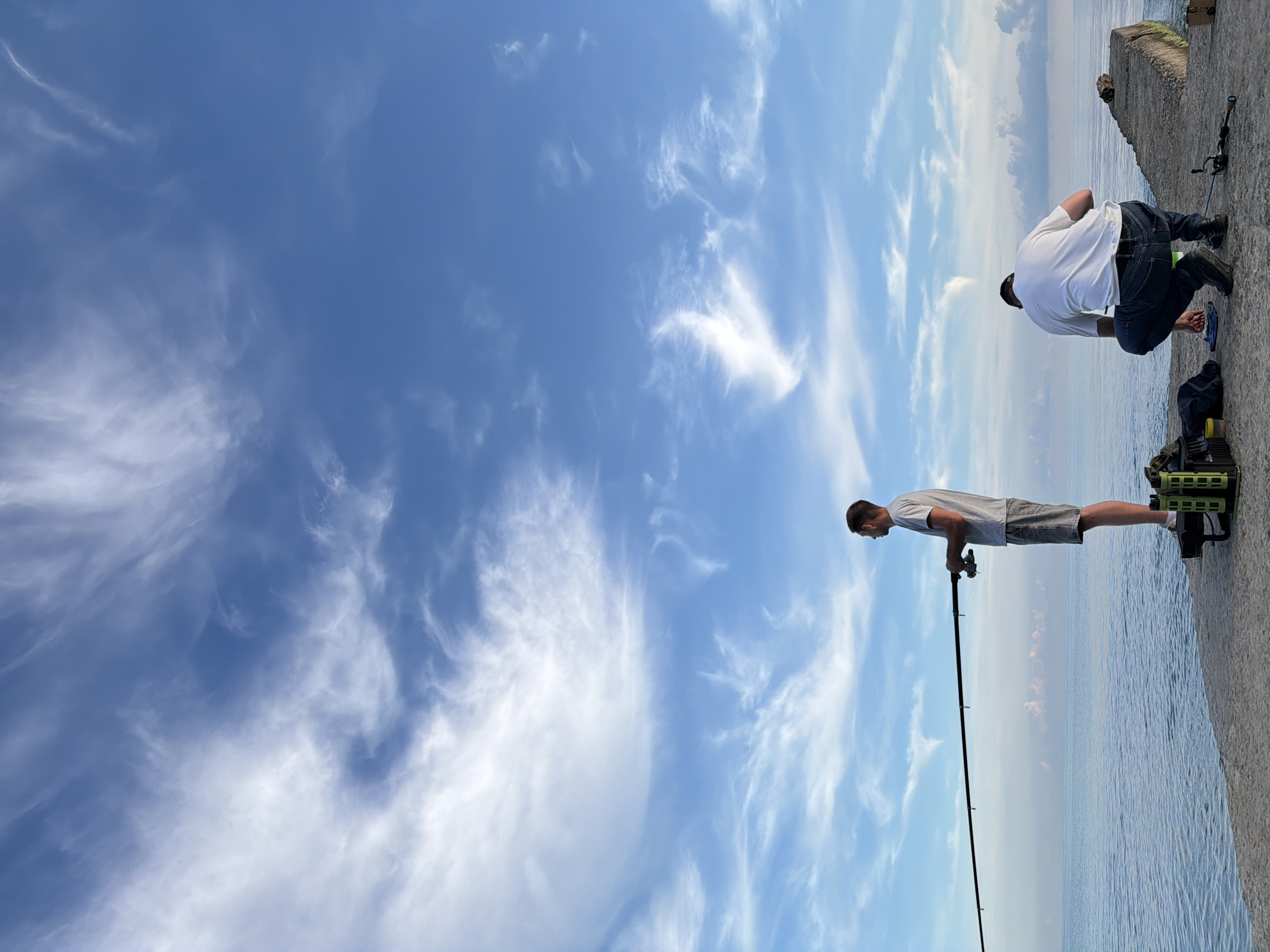 Two men are fishing on a concrete pier under a bright blue sky with wispy clouds. The man on the left stands facing the water, holding a fishing rod. He wears a gray t-shirt and denim shorts. To his right, a green fishing tackle box is visible. The man on the right crouches facing away from the camera, preparing his fishing line. He is dressed in a white t-shirt and blue jeans. A fishing rod lies on the pier near him. The sea stretches out to the horizon, with calm water reflecting the sky. The scene is set in Dederkoy, Russia.