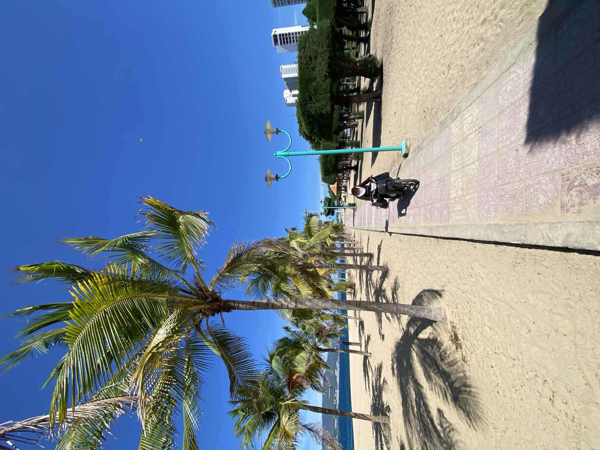 A person rides a scooter along a paved pathway next to a sandy beach lined with palm trees, in Nha Trang, Vietnam. The sky is clear and blue, and the sun is bright, casting shadows from the palm trees onto the sand. In the background, there are several tall buildings and more trees. The person on the scooter is wearing a headscarf and dark clothing, and they are carrying a bag on the front of the scooter. The pathway is decorated with patterned tiles, and there is a distinctive teal lamppost on the right side of the pathway. The scene depicts a sunny day at a tropical beach location.
