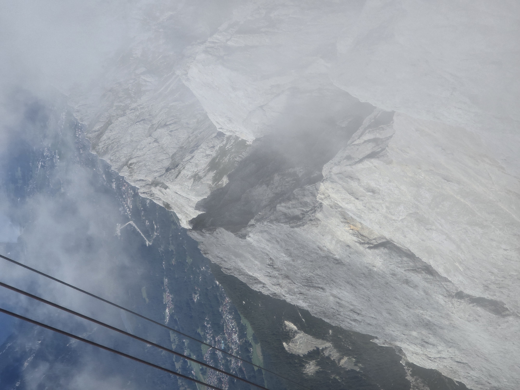 An aerial view shows a dramatic mountain range, partially obscured by clouds, with a cable car line in the foreground. The rugged, gray peaks descend into a valley below, where a small town with red-tiled roofs is nestled amongst dark green pine forests. The cable car cables, made of dark metal, stretch diagonally across the upper left portion of the image, hinting at the ascent or descent from a high vantage point. The town is situated on the slopes leading up to the mountains, with roads winding through the buildings and green fields. The overall impression is one of breathtaking natural beauty and human presence in a remote, mountainous landscape.