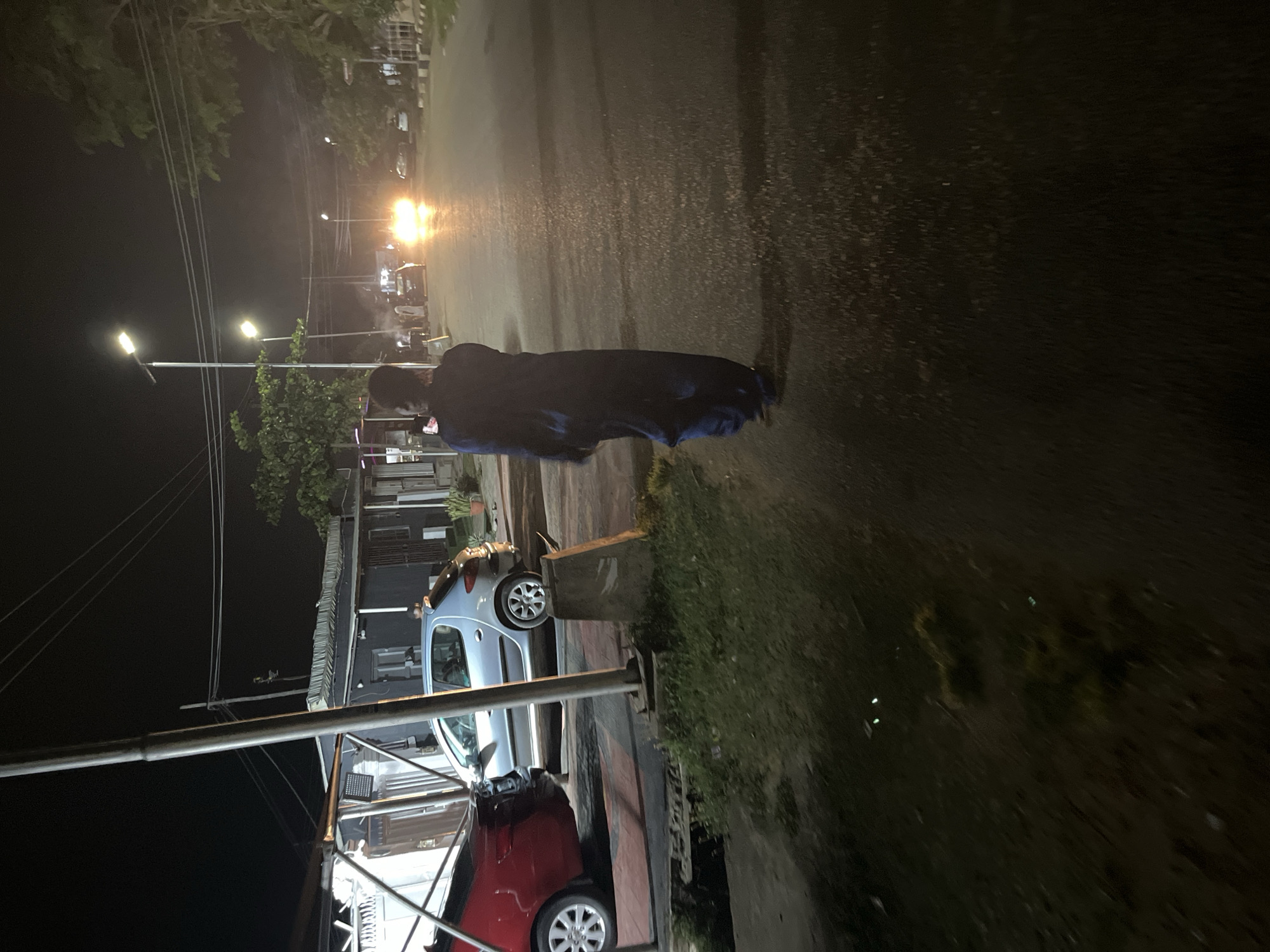 The image depicts a night scene on a street in Abuja, Nigeria. A person, seen from behind, is walking away from the camera towards the center of the frame, dressed in a dark blue robe. The street is wet, reflecting the light from multiple street lamps and the headlights of oncoming vehicles. In the background, cars are visible on the road, and buildings line the street. The environment appears to be an urban setting with a mix of commercial and residential structures. The lighting suggests it is after dark, and the presence of streetlights and vehicle headlights indicates an active street.