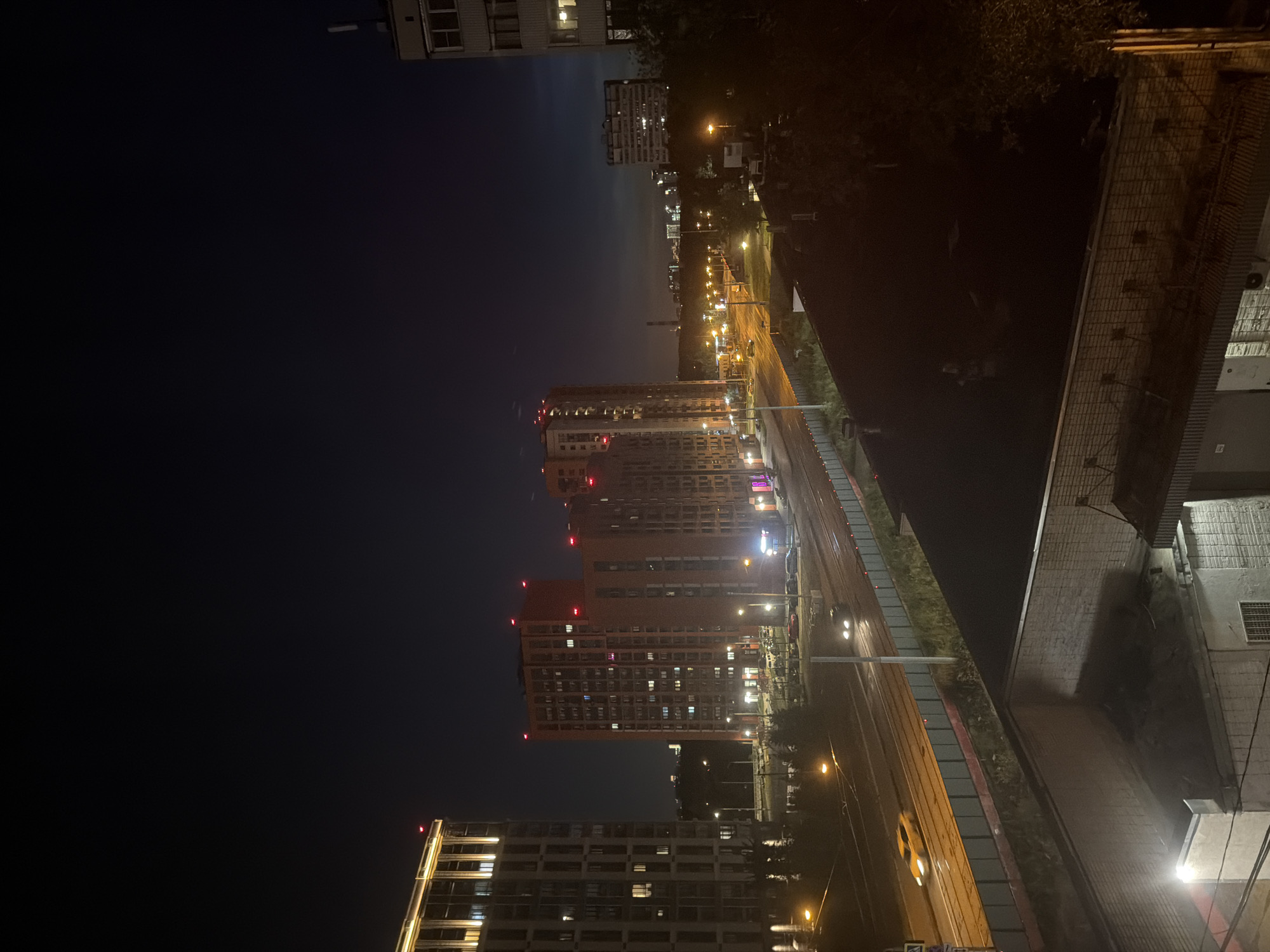 The image captures a nighttime urban scene in Moscow, Russia, likely depicting a residential area with multi-story apartment buildings and a highway. In the foreground, the dark roofs of lower buildings are visible. The mid-ground features a wide highway with active traffic, indicated by headlights and taillights of moving vehicles, including a prominent yellow car. Streetlights illuminate the roadway and the surrounding grassy areas. Several tall, modern apartment buildings with lit windows are situated beyond the highway, some featuring reddish-orange facades and red lights on their roofs. Further in the distance, more buildings and industrial structures are visible, with lights suggesting a broader urban landscape. The sky is dark with a faint gradient of lighter shades near the horizon, indicating twilight or a light-polluted sky. The overall impression is of a bustling, illuminated city at night.