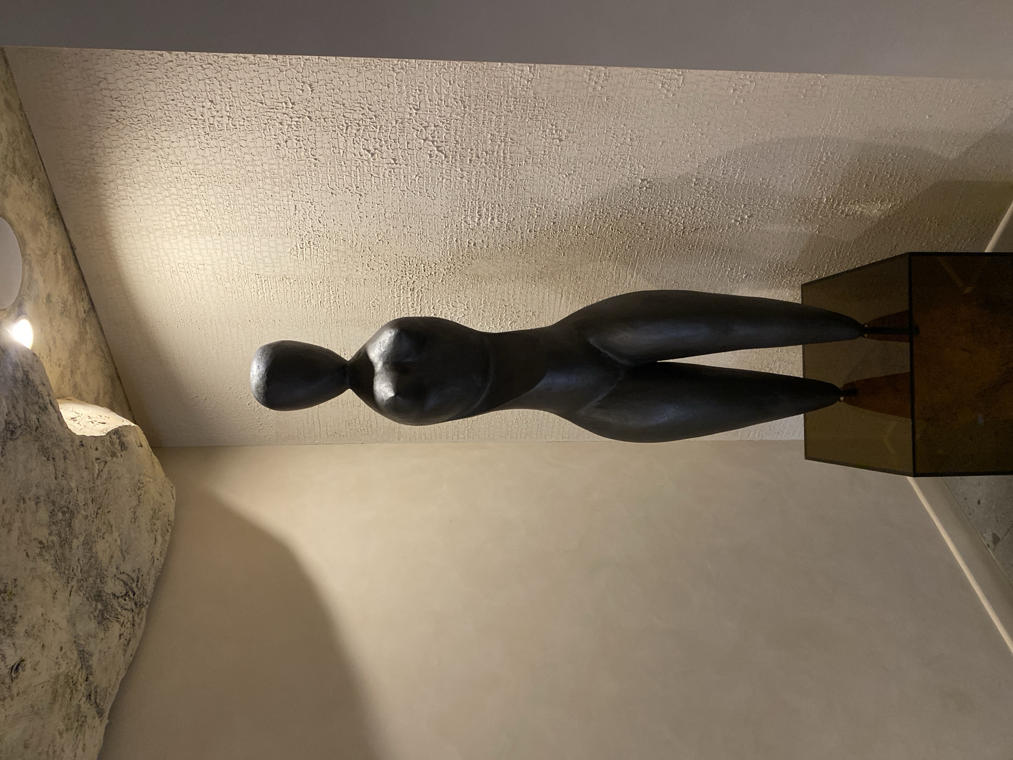 A dimly lit indoor scene features a bronze sculpture of a reclining nude female figure. The sculpture is displayed on a dark brown, possibly acrylic, pedestal. The background includes a textured ceiling, a plain beige wall, and a section of a rough, light-colored wall on the left. The location is not determinable from the image.