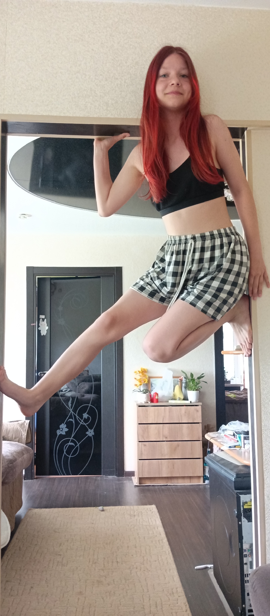 A full shot captures a female with long, red hair in a black sports bra and checkered shorts, posing in an architectural opening. She grips the upper frame with her left hand and the side frame with her right, her bare feet extended. Behind her, a door with an abstract floral design is visible. To the right, a wooden dresser is decorated with flowers and fruit. A partial view of a computer tower is seen on the right edge of the frame, resting on a hardwood floor covered by a beige patterned rug. The setting appears to be an interior space, possibly a residence. The location is described as Unknown, Unknown.