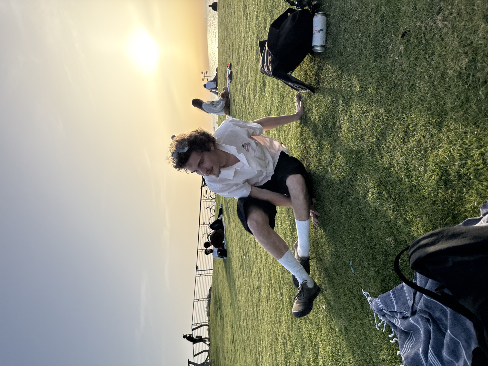 The image depicts a grassy area in Tel Aviv, Israel, likely a park or a coastal area, as the sea is visible in the background. A young man is in the foreground, crouched on the grass, wearing a light-colored short-sleeved shirt, dark shorts, white socks, and dark shoes. He appears to be adjusting his position or stretching.
In the background, several other individuals are visible, some seated or lounging on the grass. A metal railing is also present, indicating a boundary or edge of the area. A black backpack and a can of "San Miguel" beer are positioned on the grass near the man. The sun is setting, casting a warm, golden light across the scene.