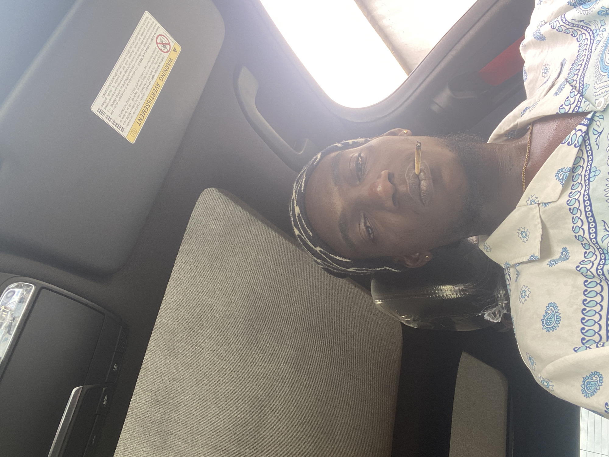 A man is seated in the driver's seat of a car, looking directly at the camera. He is wearing a bandana and a white shirt with a blue paisley pattern. A lit cigarette is in his mouth. The interior of the car is visible, including the sunroof and a warning sticker on the sun visor. The scene appears to be in Oniyangi, Nigeria, though there is no specific visual information in the image to confirm this location. The lighting suggests it is daytime.