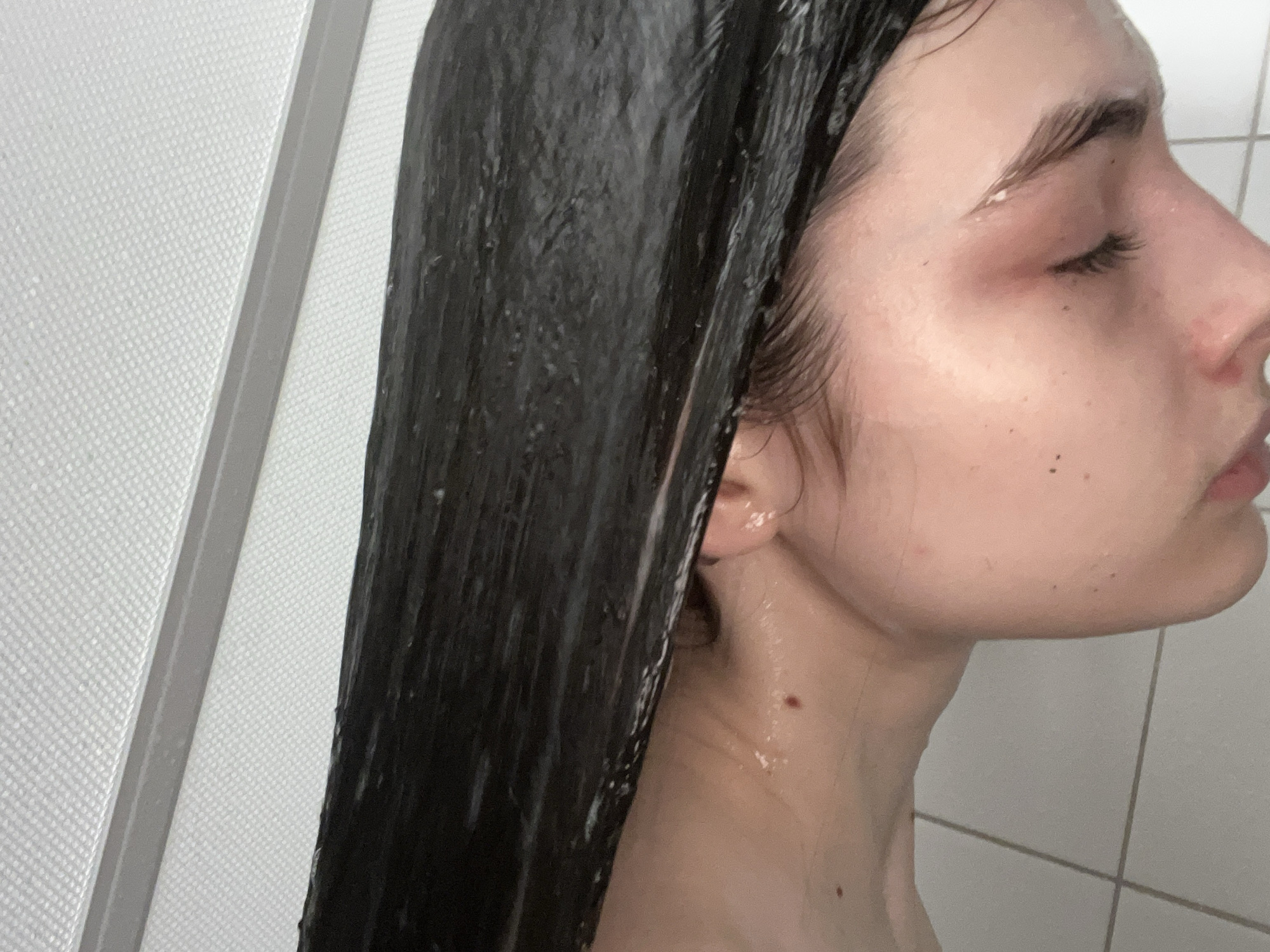 A close-up shot of a woman with closed eyes, wet, dark hair, and fair skin, appearing to be in a shower. The background shows a white tiled wall. The city and country are undetermined.