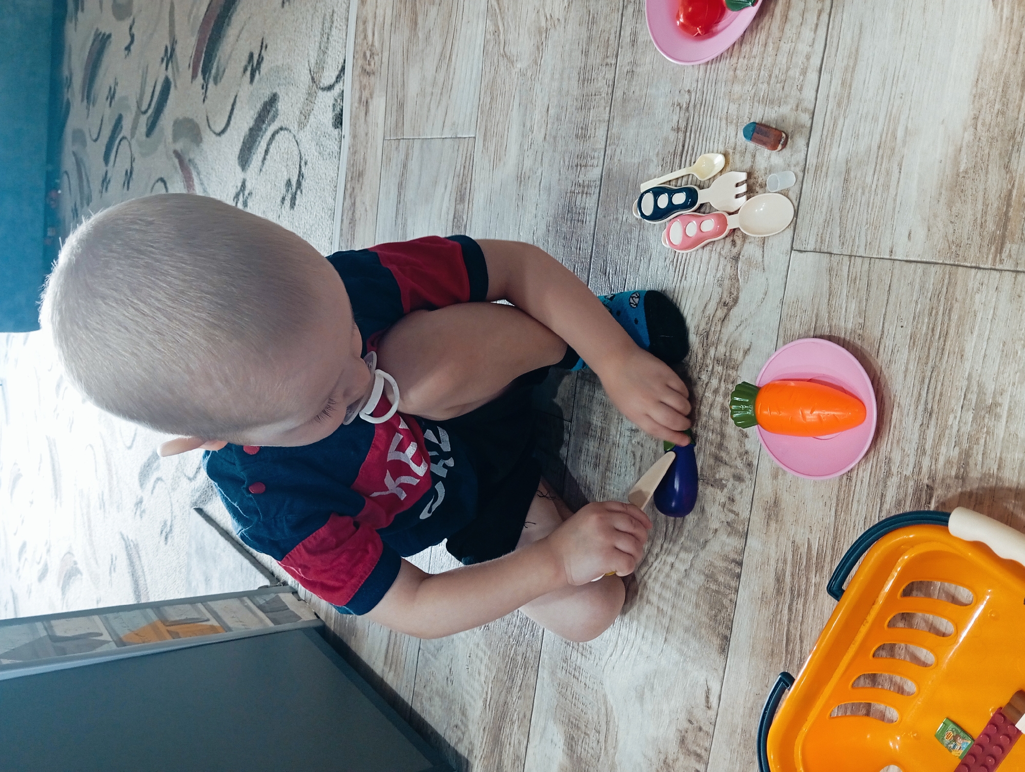A child is sitting on a floor with wood-grain-patterned flooring, playing with toy food items. The child, who appears to be a toddler with fair hair, is wearing a short-sleeved shirt with a red upper portion and a dark blue lower portion, along with dark shorts. A pacifier is in the child's mouth. The child is holding a toy eggplant and a toy knife. Around the child are various toy food items and utensils, including a toy carrot on a pink plate, a toy tomato on another pink plate, toy spoons and forks, and a small block. An orange toy shopping basket is partially visible on the right side of the image. The flooring shows a pattern of wood grain with some areas that appear lighter and more worn. The room's walls have a pale patterned design. The location is Yurgamysh, Russia.