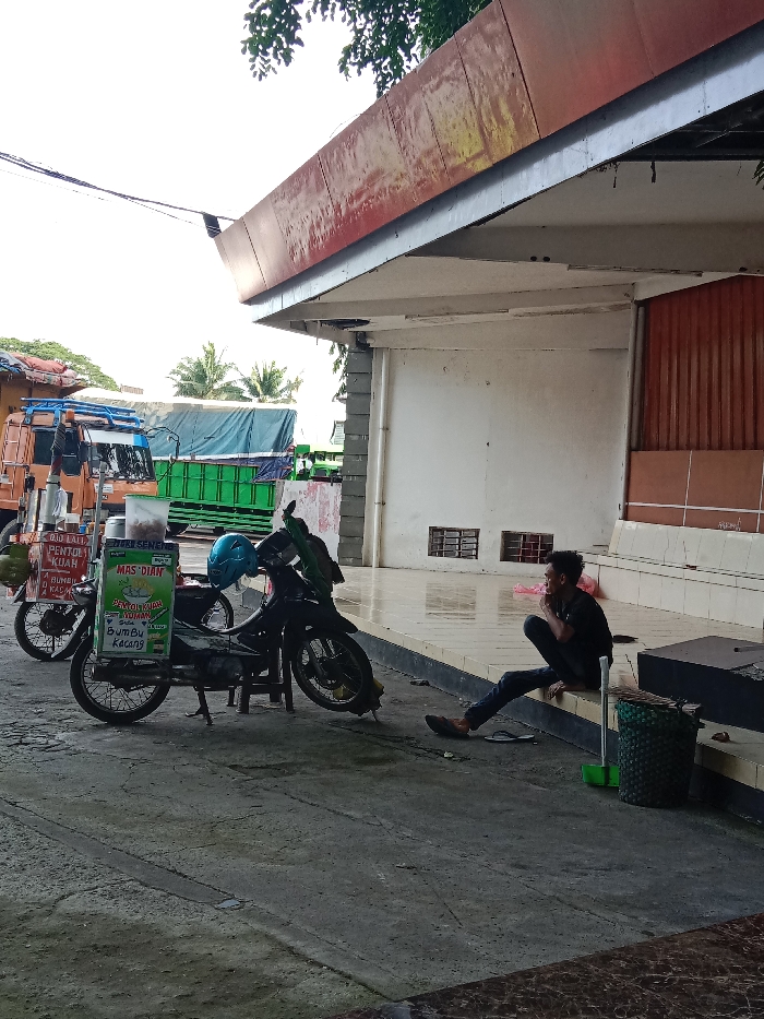 The image displays an outdoor scene in Wonoketingal, Indonesia, under daylight conditions. A single male individual, wearing a dark shirt and pants, is seated on the edge of a raised, light-colored tiled platform located on the right side of the frame. One dark sandal is on his foot, and another is on the ground beside him. His right hand is positioned near his mouth.

To the left of the seated man, two dark-colored motorcycles, adapted as mobile vending stalls, are parked on a gray concrete surface. The motorcycle on the left features a side-mounted stall advertising "PENTOL KUAH" and "BUMBU KACANG," with a green coconut suspended from its front. The motorcycle on the right has a prominent green vertical sign displaying "MAS DIAN" and "PENTOL KUAH ISTIMEWA BUMBU KACANG"; a light blue helmet rests on its seat.

In the background, multiple commercial trucks are visible, including an orange truck with a blue tarp-covered load and several green trucks. The area is partially covered by a weathered red-orange corrugated metal canopy, supported by concrete pillars, extending from a building with a white wall and brown corrugated metal paneling. Two rectangular vents are present in the white wall. A green broom and a dark green woven basket are on the ground adjacent to the seated man. Overhead power lines traverse the upper left portion of the frame against a bright sky.