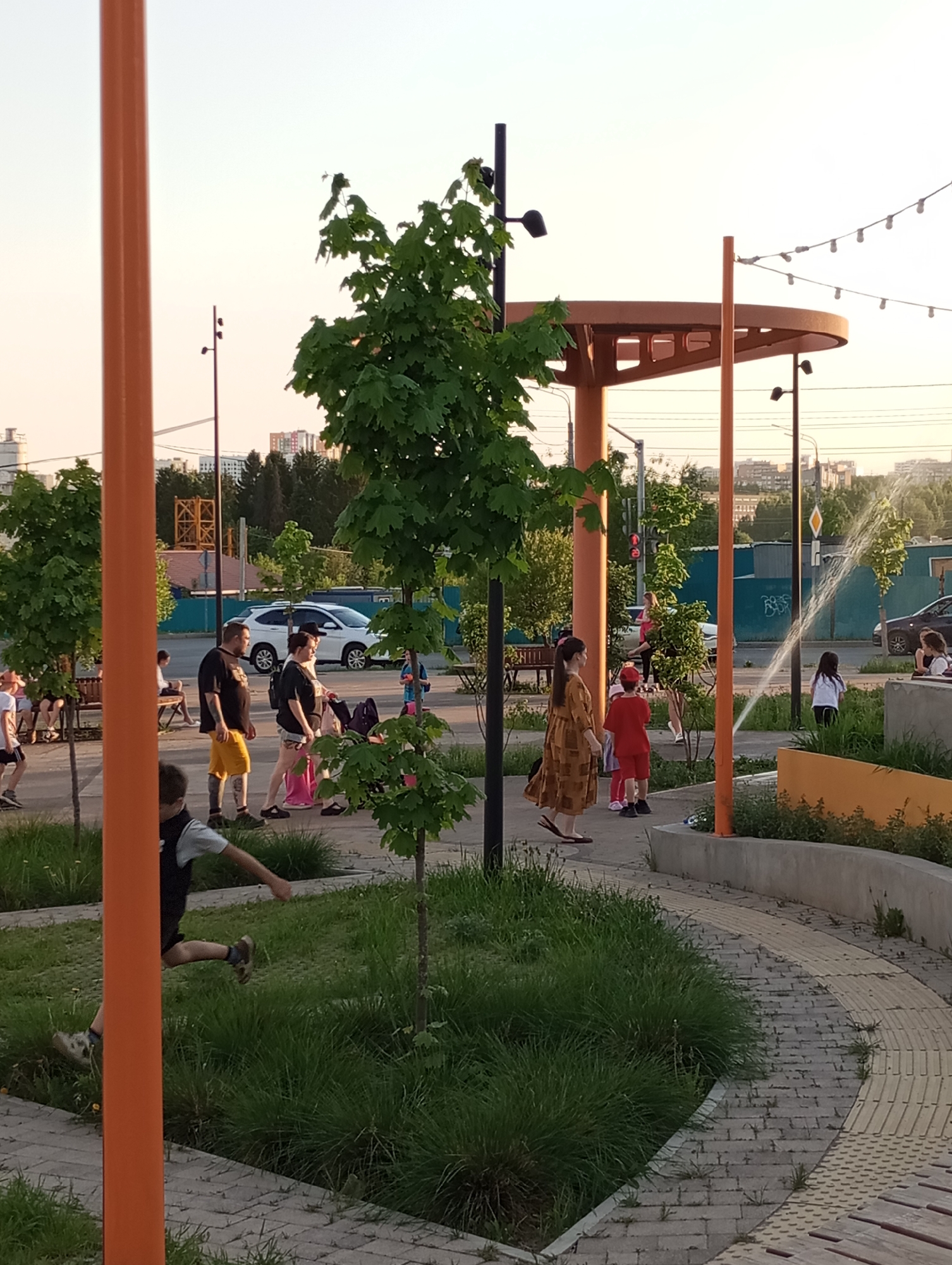 The image depicts a public park in Pervomaysky, Russia, during daytime. A paved path curves through a grassy area. A young boy wearing a dark vest is jumping. People are strolling in the background. Features include trees, orange poles, a structure providing shade, and a water fountain feature. Other individuals are also walking around. Buildings are seen in the background.