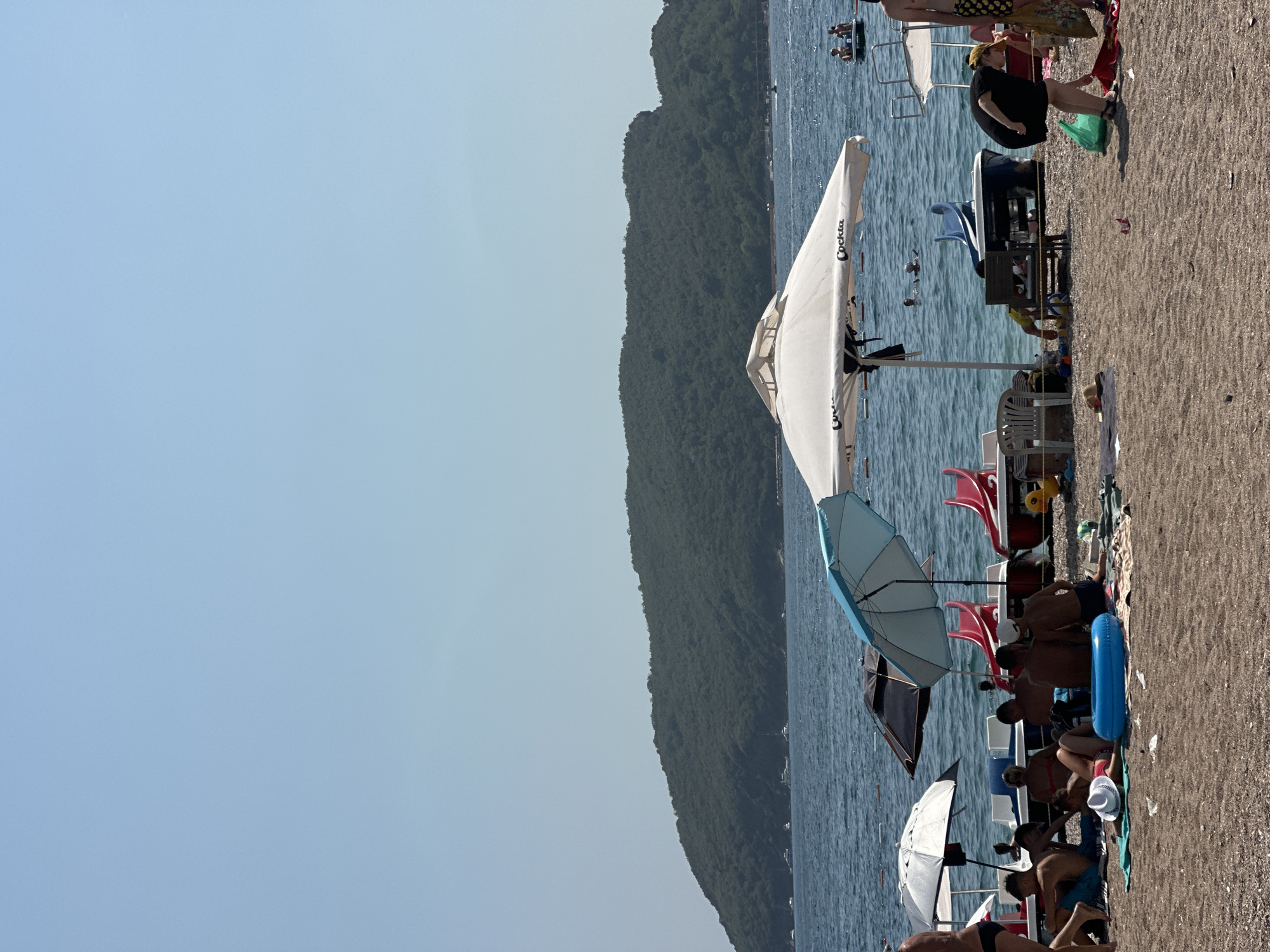 The image depicts a sunny day at a beach in Budva, Montenegro. The foreground is a sandy beach with several groups of people relaxing under beach umbrellas and on towels. Some individuals are seated, while others are lying down. A blue inflatable ring is visible on the sand.
In the mid-ground, the blue sea stretches out, dotted with several boats and pedal boats. The water is calm, and a few people can be seen swimming. Along the shoreline, there are more beach umbrellas and beach chairs.
In the background, a large, green, tree-covered hill dominates the landscape, likely a characteristic feature of the Budva coastline. The sky is a clear, pale blue. The overall scene conveys a typical summer beach atmosphere with people enjoying leisure activities by the sea.
