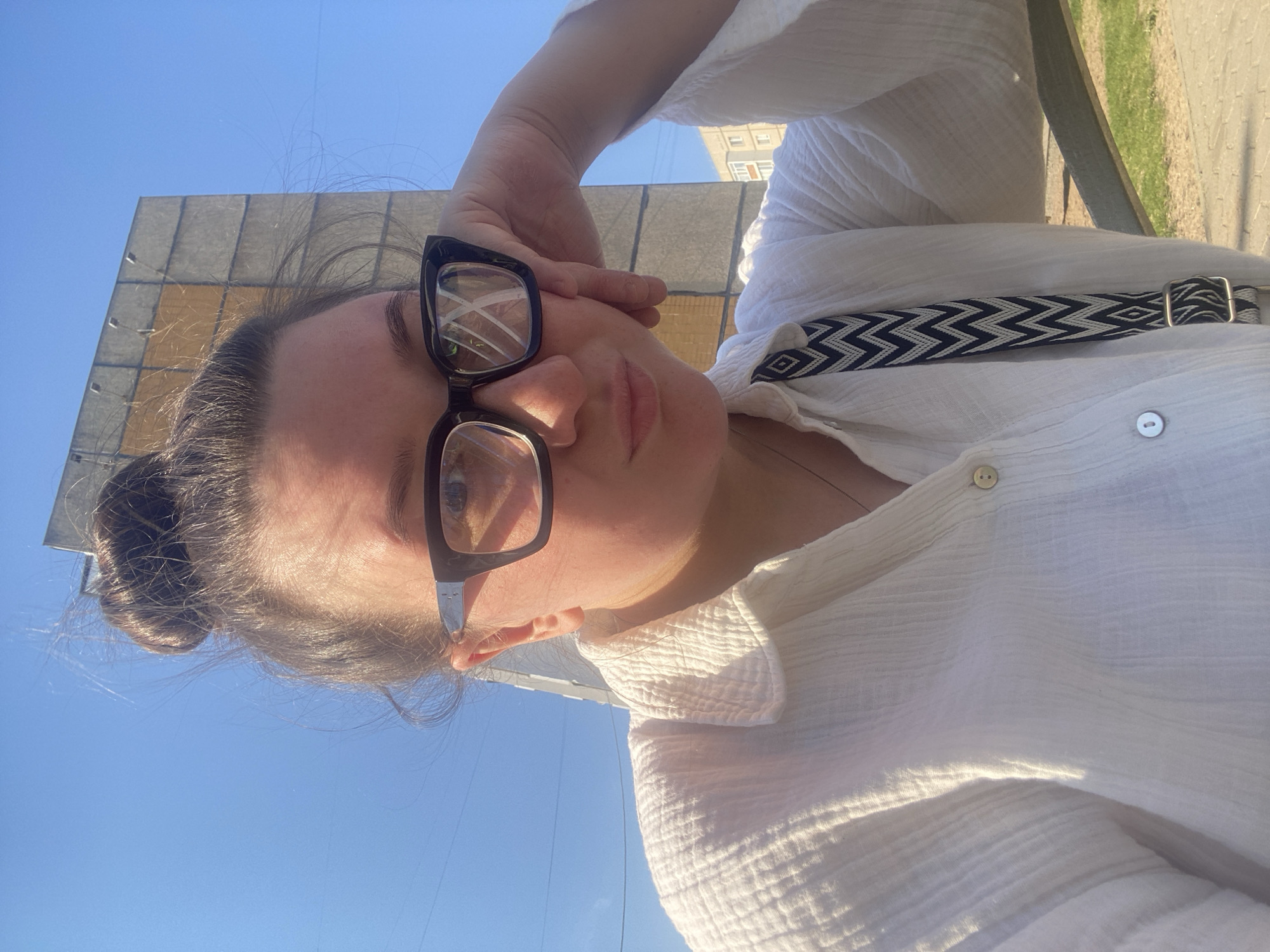 A woman is seen in a selfie-style photograph. She is wearing black-framed glasses and a white, textured button-down shirt with a black and white patterned strap running across her chest. Her hair is pulled back into a bun. Behind her, there is a light blue sky and a light brown rectangular building. Based on the background, the photo was taken in an urban area. Based on the description provided by the user, the location of this picture is Chaikovsky, Russia.