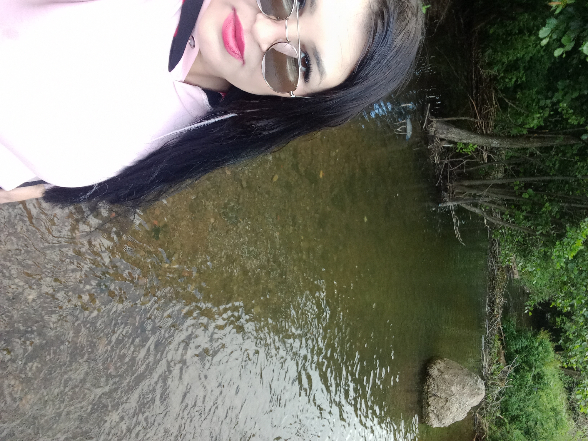 The media file is a self-portrait of a woman taken outdoors. The woman is wearing sunglasses and a pink top with a black collar. She has long black hair and bright pink lipstick. She is standing in shallow water, possibly a river or stream, with trees and foliage visible in the background. A large rock is submerged in the water to her right. The lighting suggests it is daytime. The overall mood appears casual and relaxed. There is no visible text in the media file.