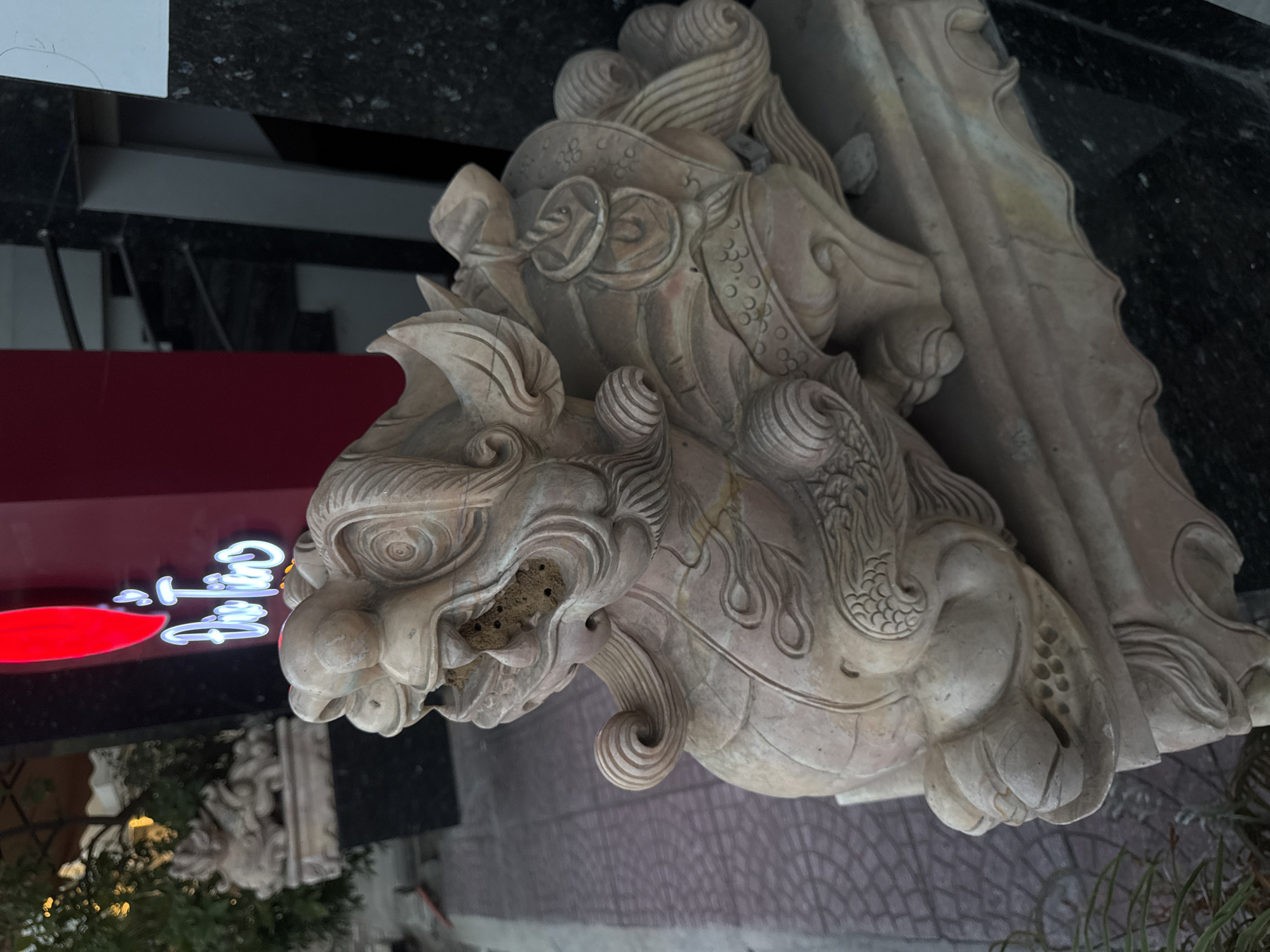 The image displays a close-up, eye-level shot of a stone sculpture of a Foo dog, a mythical creature often used as a guardian in East Asian cultures. The sculpture is light brown with visible cracks and weathering, suggesting it is an outdoor fixture. It is seated on a decorative stone base. In the background, a red sign with white neon lettering, which reads "Đức Tân," is visible. The lettering appears to be in Vietnamese. To the left of the sign, out-of-focus greenery can be seen, along with another, less distinct Foo dog sculpture. The ground in front of the sculpture is paved with square tiles. The scene suggests the sculpture is located outside a business establishment in Da Nang, Vietnam, likely for decorative or protective purposes.