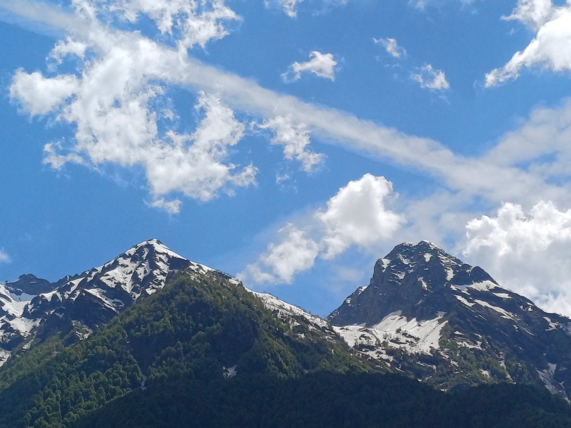 This landscape image captures a majestic view of snow-capped mountains under a vibrant blue sky. Two rugged mountain peaks dominate the lower half of the frame, showcasing rocky terrain with significant patches of white snow, particularly near their summits. Below the snowline, the mountains are densely covered in dark green coniferous forests, extending down towards the foreground.
The sky is a brilliant azure, adorned with scattered, fluffy white cumulus clouds of various shapes and sizes. A distinct, wispy white cloud or contrail stretches diagonally across the upper right section of the sky, adding a linear element to the cloudscape. The scene is bathed in bright daylight, indicating a sunny day, with clear distinctions between light and shadow on the mountain surfaces, highlighting their contours.
No people, man-made objects, or visible text are present in the image. The natural setting evokes a sense of grandeur, peace, and the raw beauty of a mountainous region. The presence of both snow and lush green forests suggests a season like late spring or early summer in a high-altitude location. The provided context indicates this scenic view is located in Estosadok, Russia.
