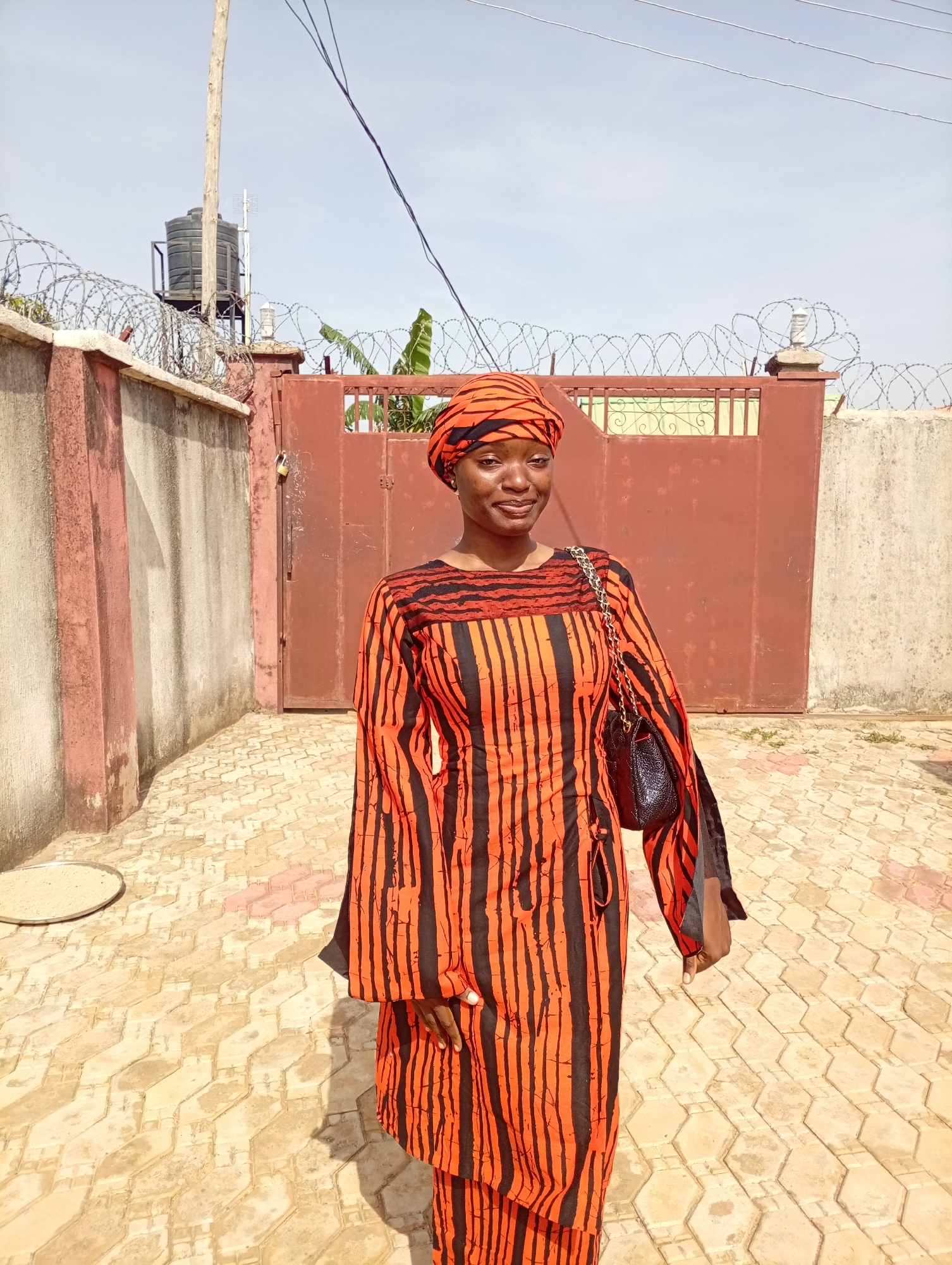 A young woman is standing in a courtyard of a building in Unknown, Unknown. The woman is wearing a matching orange and black striped dress and head covering. She is also wearing a black purse with a chain strap. The courtyard is paved with hexagonal stones. In the background, there is a red metal gate with barbed wire on top, and a concrete wall with a water tank on top.