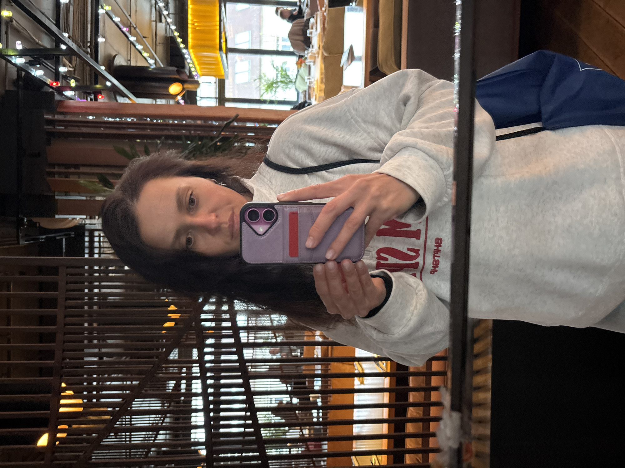 An indoor mirror selfie captures a woman in a casual setting, likely a cafe or restaurant in Kazan, Russia. The central subject is a woman with long, dark hair, wearing a light gray sweatshirt with red, reversed text on it (partially visible as "2I8THB"). She holds a purple smartphone with a red stripe, looking directly into the reflective surface.
The background, visible through reflection, shows an eclectic interior with warm yellow lights, string lights, dark metal beams, and wooden slatted structures that resemble blinds or decorative panels. Through what appears to be a large window, an exterior building with multiple windows is reflected, suggesting daylight outside. Hints of other patrons and a plant are also visible in the reflections, contributing to the ambiance of a public space. The image conveys a contemporary urban scene, captured casually.