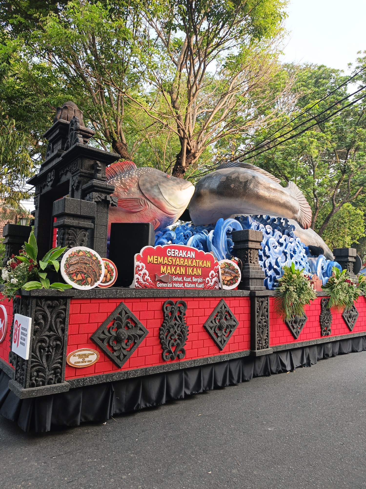 This image captures a parade float in the city of Gayamprit, Indonesia, likely during a promotional event. The float is adorned with large, stylized fish figures, one predominantly gray and the other with red fins. They are positioned above a vibrant red facade decorated with black, ornate patterns resembling traditional Indonesian motifs. The float also features representations of blue waves, adding a dynamic element to the display. In front of the fish, two plates of what appears to be cooked fish dishes are prominently displayed on white platters. A red sign with white text, partially obscured by the fish, reads "GERAKAN MEMASYARAKATKAN MAKAN IKAN" which translates to "Movement to Popularize Eating Fish." Below this, smaller text states "Sehat, Kuat, Bergizi - Generasi Hebat, Klaten Kuat!", meaning "Healthy, Strong, Nutritious - Great Generation, Strong Klaten!". The float is decorated with floral arrangements and has a black, ruffled skirting around its base. In the background, lush green trees and a bright sky suggest an outdoor setting on a sunny day. The float is situated on an asphalt road, indicating it is part of a procession.