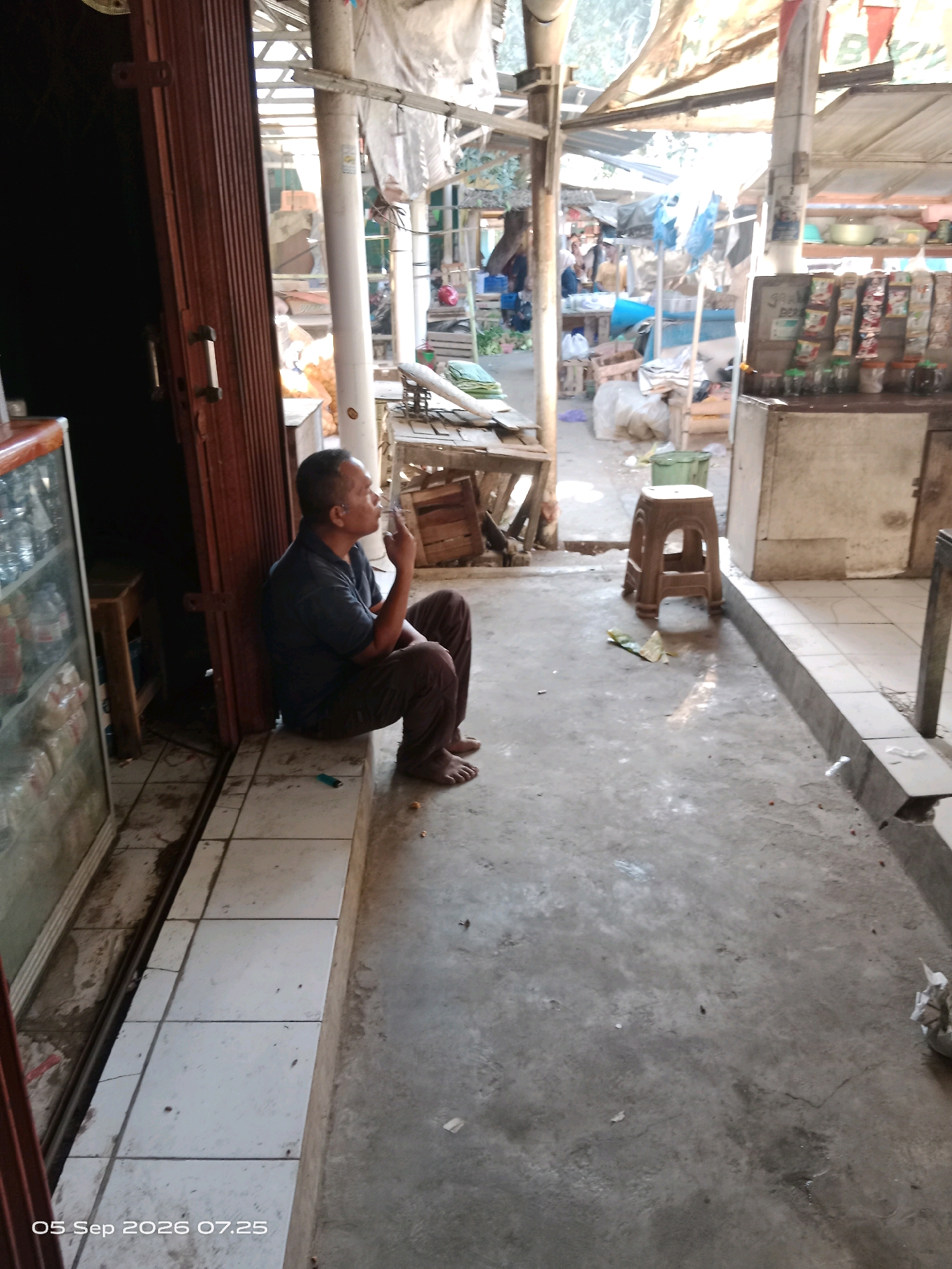 In Kalipurwo, Indonesia, a man sits on a tiled ledge in front of a shop. He is wearing a dark blue t-shirt and dark pants, and appears to be smoking a cigarette. The shop has a wooden door and a display cooler to the left. The surrounding area is an outdoor market or street scene with stalls and various items visible in the background. The ground is concrete, with some debris scattered around.