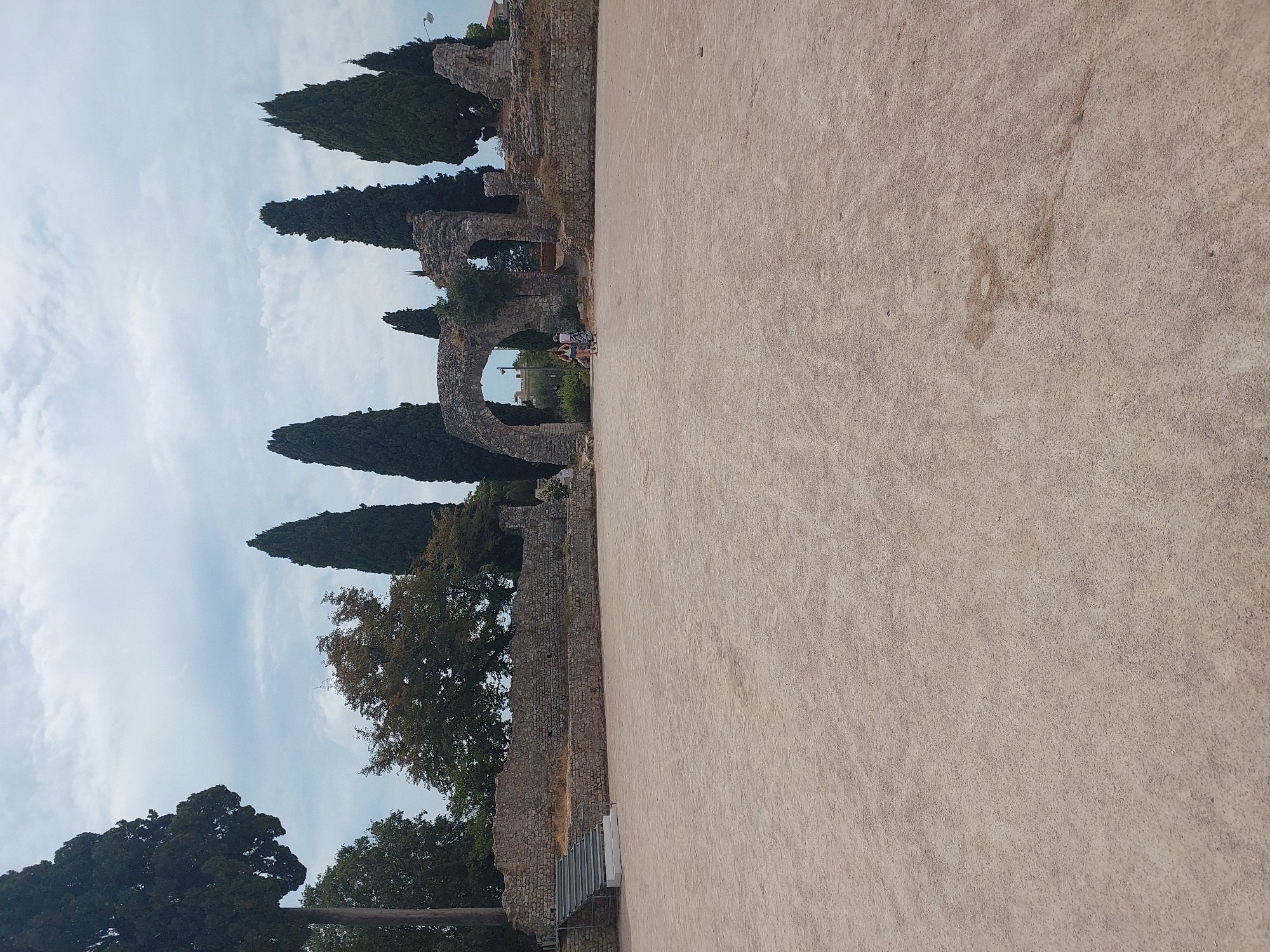 The image shows an outdoor archaeological site in Nice, France, featuring ancient stone ruins and a large, open sandy area. Tall, slender cypress trees are prominent around the ruins, and a few deciduous trees are also visible. The sky is overcast with clouds. In the mid-ground, there is a large arched stone structure, part of the ruins, with two individuals walking through it. To the left of the ruins, a modern metal staircase with white steps leads up to a stone wall. The foreground is a flat expanse of light-colored sand or gravel. The overall impression is of an historical site under a cloudy sky.