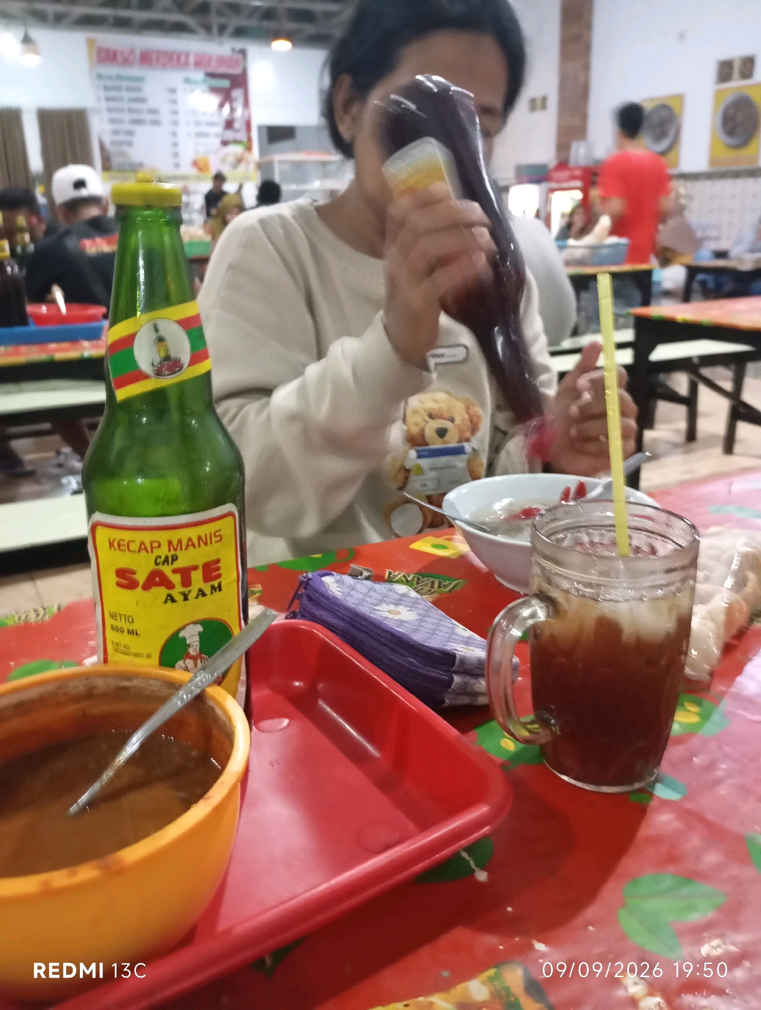 The image depicts a table setting at a restaurant in Wuluhan, Indonesia, likely a local eatery given the signage in Indonesian. In the foreground, a green bottle of "Kecap Manis Cap Sate Ayam" (sweet soy sauce for chicken satay) stands prominently. Beside it, a red tray holds a small orange bowl with a spoon and a serving of a dark sauce, presumably related to satay. To the right, a glass mug filled with a dark beverage, likely iced tea, is visible with a yellow straw. A stack of purple-patterned napkins sits between the soy sauce bottle and the beverage. In the background, a person is partially visible, holding a squeeze bottle, seemingly adding sauce to food, though the food itself is out of frame. Other patrons are blurred in the background, and a menu board displaying "Bakso Merdeka Wuluhan" is visible on the wall, confirming the location. The overall environment suggests a casual dining experience.