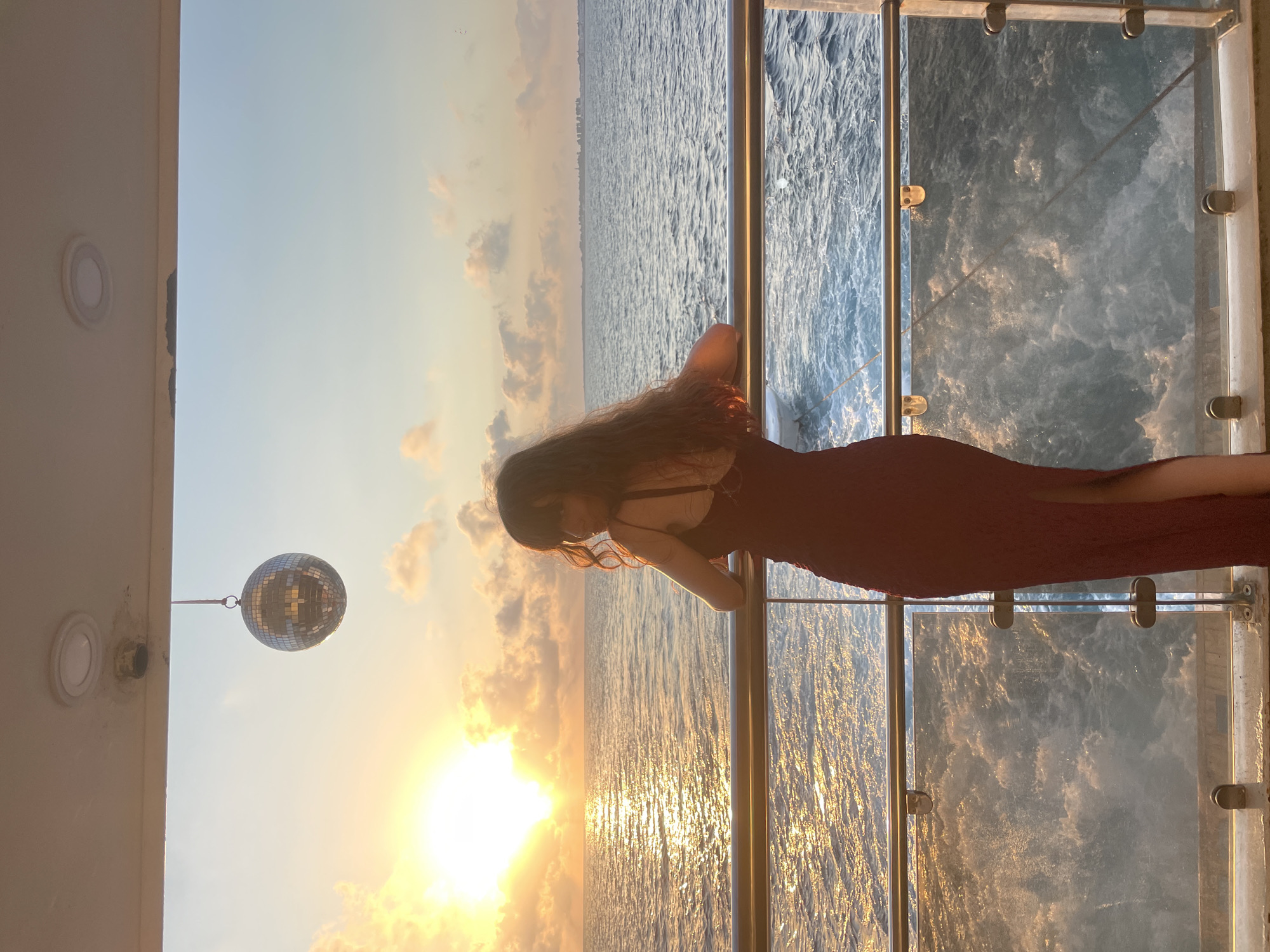 A woman with long, dark, wavy hair is standing on the deck of a boat, leaning on a railing and looking out at the sea. She is wearing a long, dark red dress with a slit up the side. The sun is setting over the water, casting a golden glow. A disco ball hangs from the ceiling above her. The boat appears to be moving, as evidenced by the wake in the water. The scene suggests a celebration or special occasion.