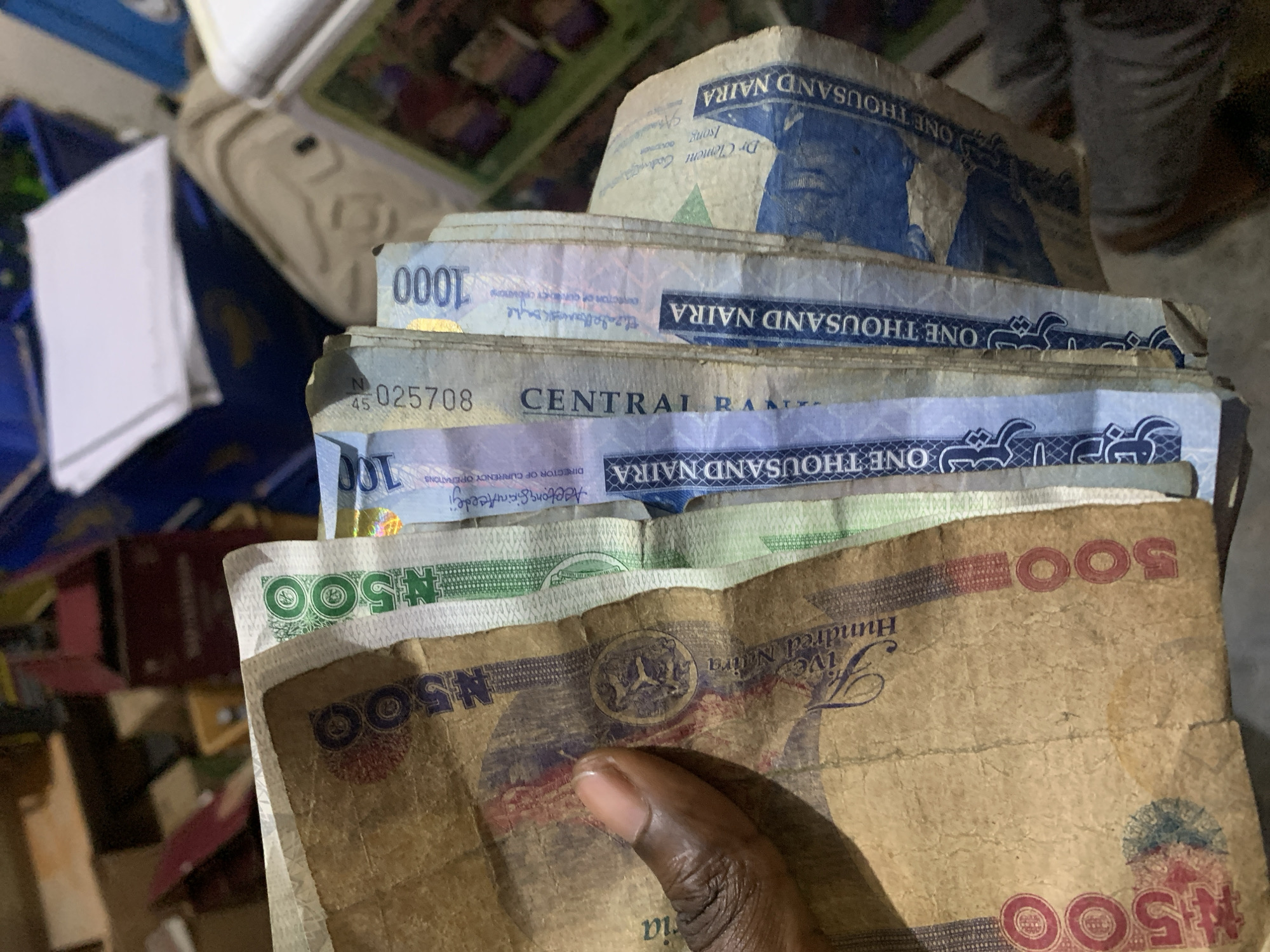 The image shows a person holding a stack of Nigerian Naira banknotes. The denominations visible include 500 Naira and 1000 Naira notes. The person's hand and part of their arm are visible in the lower part of the image. The background is blurry and includes glimpses of other objects and surfaces. There is no other location information in the images provided.