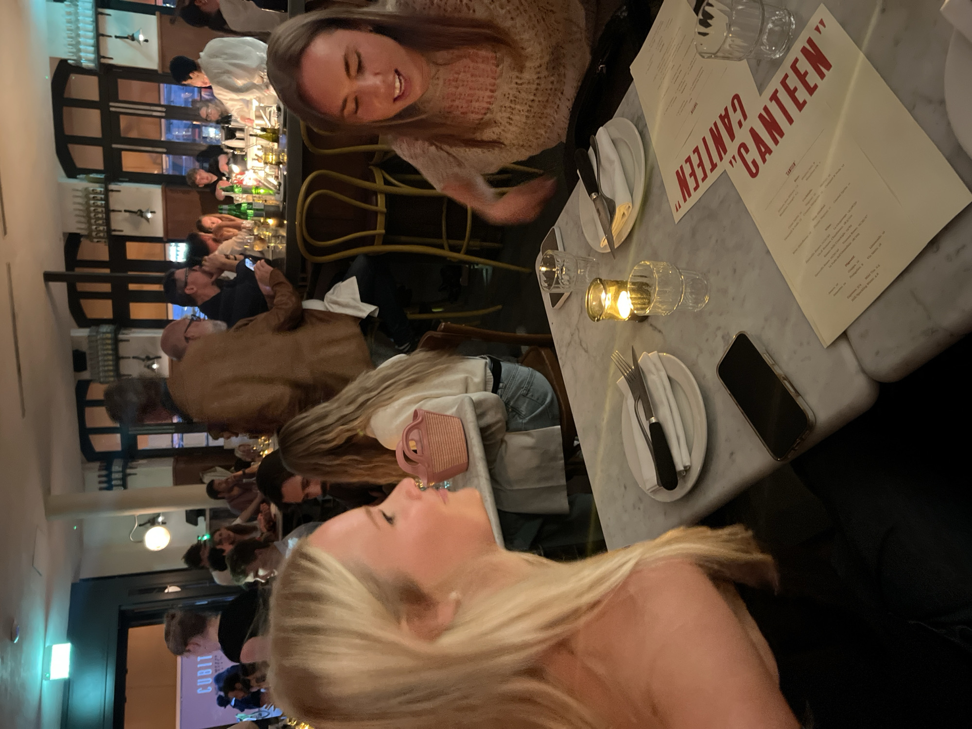 The image shows the interior of a restaurant called "Canteen," likely located in London, United Kingdom. Several people are present, including two women at the foreground of the image. One of the women has blonde hair. Another has brown hair and is smiling. A menu is visible on the table. Other details include cutlery, glassware, a mobile phone, a purse and a lit candle. There is bar seating and other patrons at the restaurant. The light fixtures and overall decor suggest a modern or upscale environment.