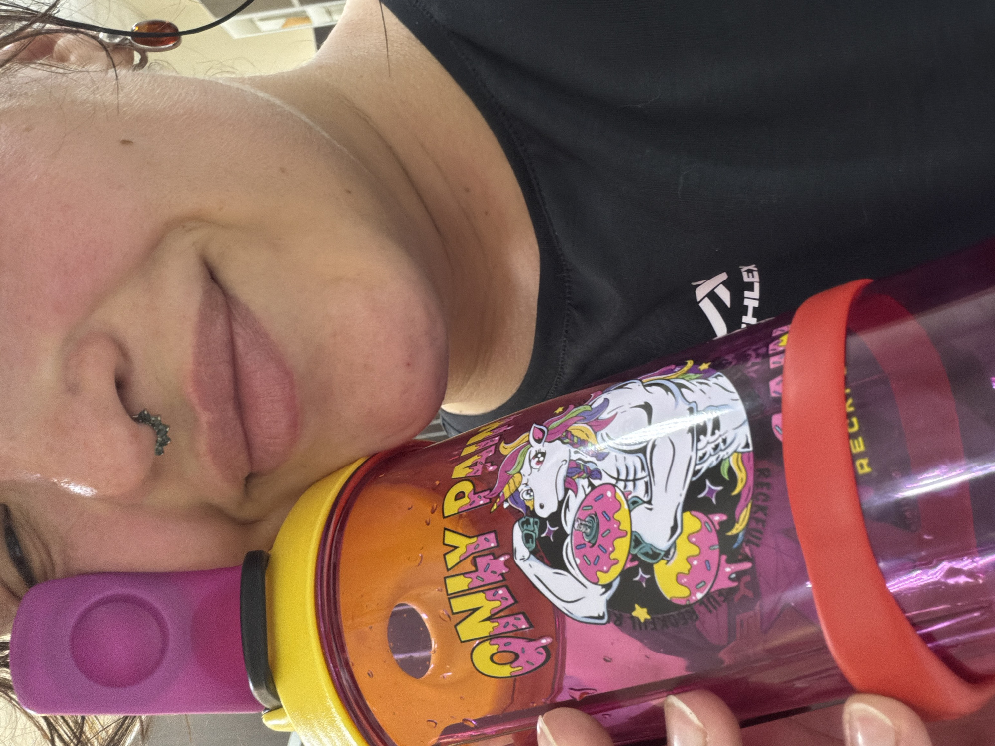 A close-up, medium shot shows a person's face and shoulder holding a pink water bottle. The person is wearing a black t-shirt with a white logo on the right side. They have a septum piercing and large orange earrings. The water bottle has a yellow cap with a purple lid and features a graphic of a muscular unicorn eating a donut, with the words "ONLY PAIN" in pink letters. The background is blurred and appears to be indoors, with a hint of white cabinetry and what looks like office lighting. The image does not provide any contextual information that would allow for identification of its location within Chaikovsky, Russia.