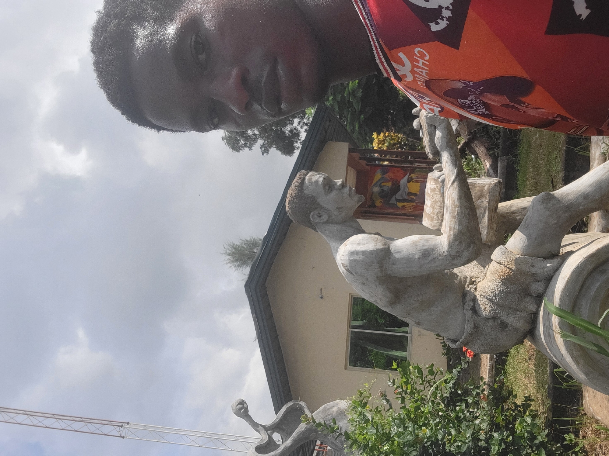 A young Black male, facing the camera and looking slightly off-center, is in the foreground of the image. He is wearing a red and orange shirt with a white and black patterned collar and graphics. Behind him, to the left, is a large, light-gray concrete sculpture of a man in a seated position, appearing to be engaged in manual labor with a wooden object. To the far left, another abstract concrete sculpture is partially visible. A building with a tan facade and a dark roof is situated behind the sculptures. The building has a window with reflections of trees. Green foliage and grass are visible around the sculptures and in front of the building. A tall metal structure, possibly a communication tower, stands in the background to the upper left. The sky is overcast with some clouds. The scene is an outdoor setting, likely a public space or garden, with hints of the environment of Egbagu, Nigeria, suggesting a cultural or artistic display.