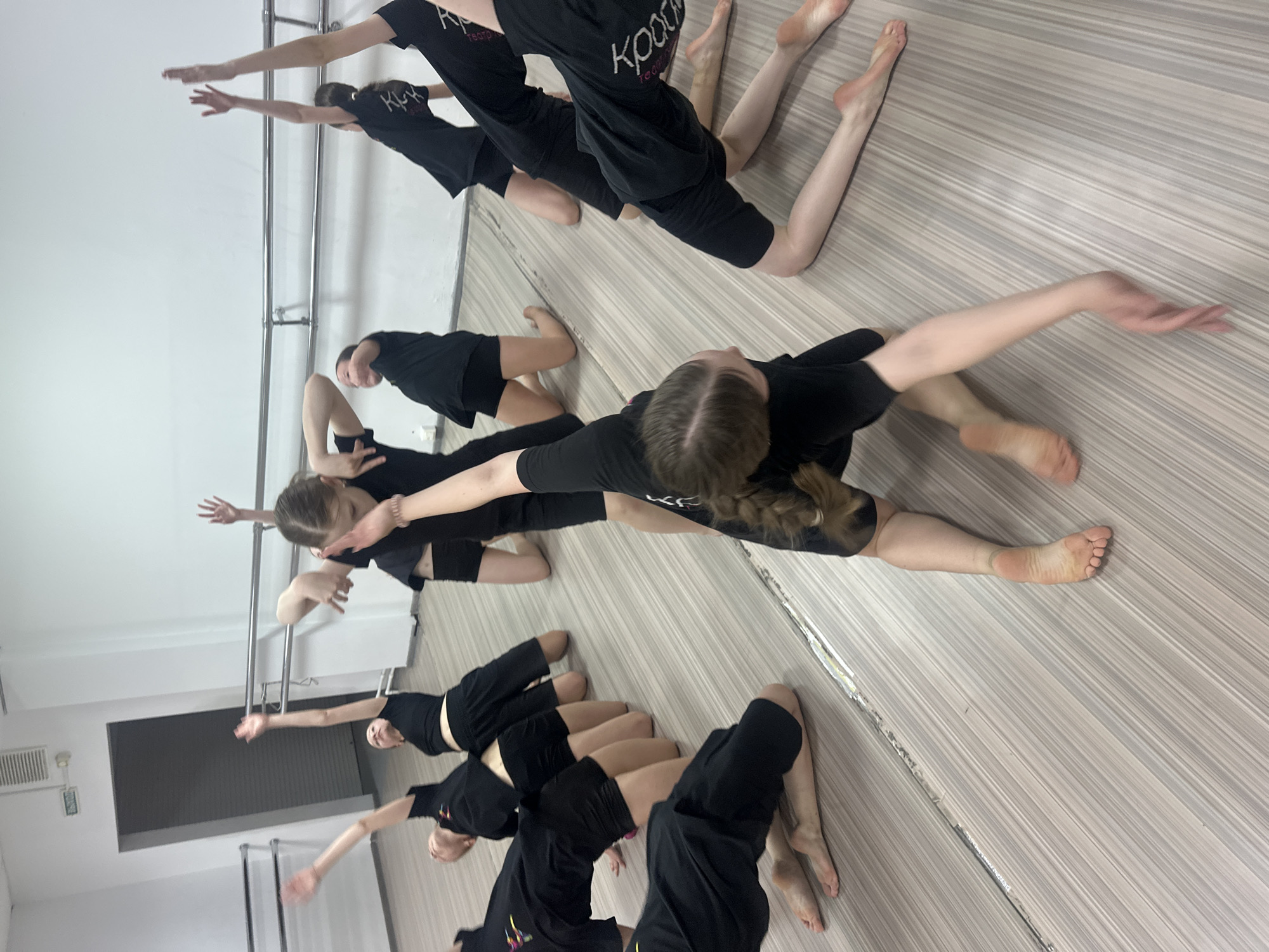 The image shows a dance practice in a mirrored studio, likely in Votkinsk, Russia. Several young dancers, all dressed in black T-shirts and shorts, are positioned on the floor. They are barefoot and in a kneeling position, with their arms extended. Some are reaching upwards towards the barre attached to the wall. The studio has a wooden floor, bright lighting, and a mirrored wall that reflects the dancers and the room, creating a sense of depth. There's a barre mounted to the wall. The girls are practicing a dance move in a synchronized manner.