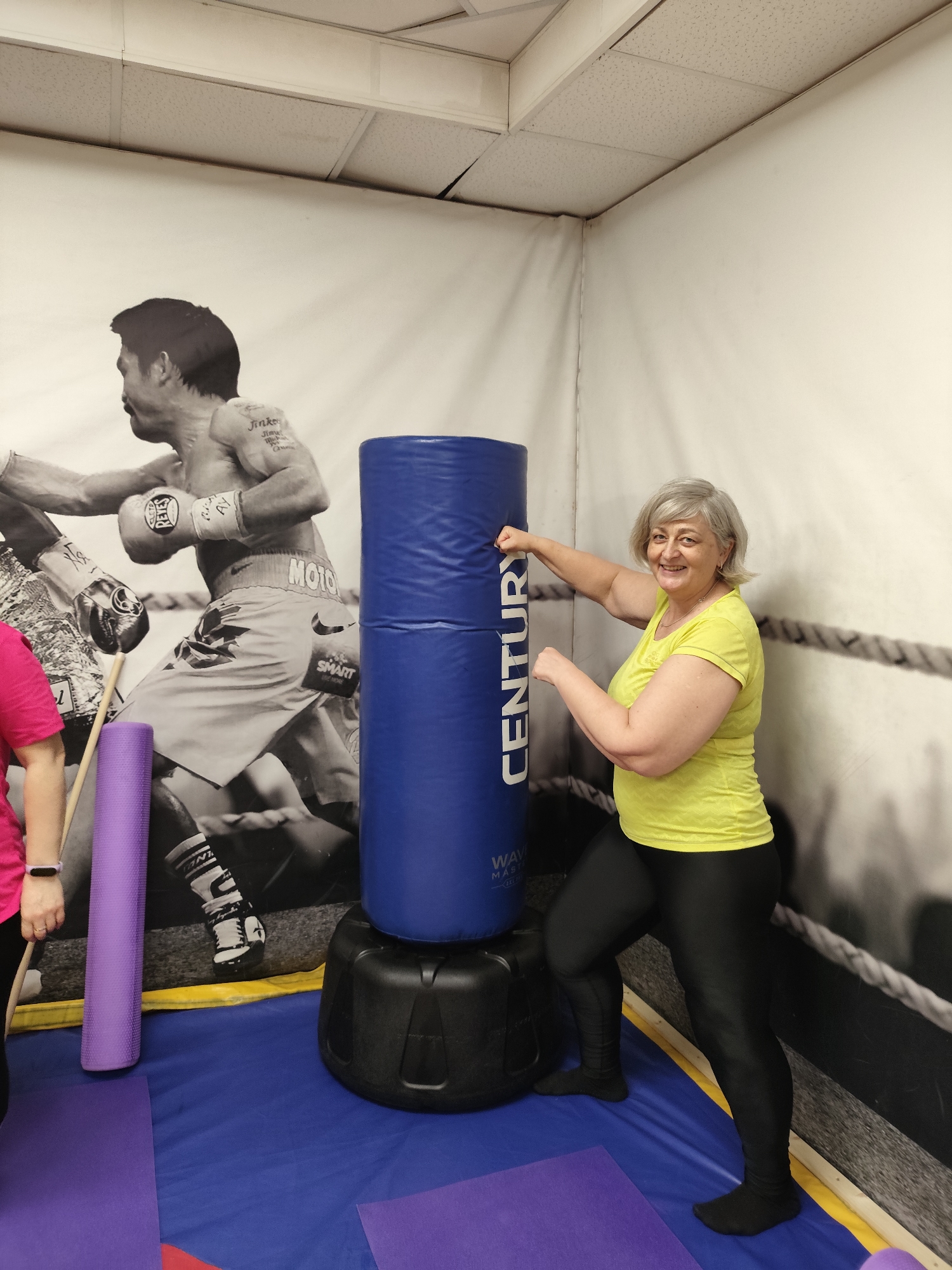 A middle-aged woman with short gray hair and a yellow t-shirt and black leggings stands in front of a large blue punching bag, with her right arm extended and fist touching the bag. She is smiling at the camera. Behind her is a large mural of a boxer. In the foreground, there are two rolled-up yoga mats, one purple and one pinkish-purple, on a blue mat. The scene appears to be a gym or fitness studio. There is no information available to determine the location in the city of Krasnodar, Russia.