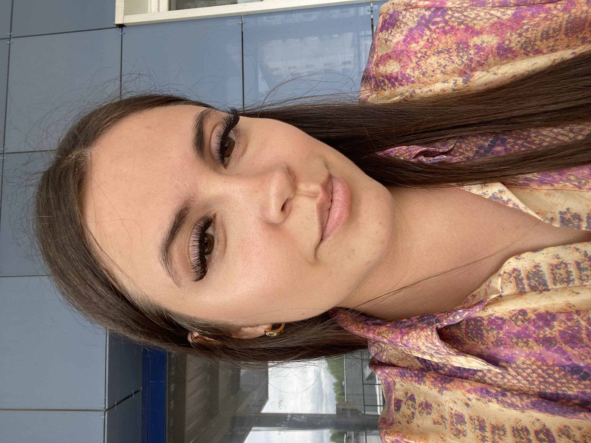 A selfie of a young woman with long, dark brown hair is taken outdoors, with a blue, tiled building and cloudy sky in the background. The woman is wearing a colorful blouse with a snakeskin-like pattern in shades of yellow, pink, and purple. She has long, dramatic false eyelashes and is wearing a small gold hoop earring. She is looking towards the camera with a slight smile. The background suggests an urban or commercial setting, possibly a balcony or outdoor seating area.