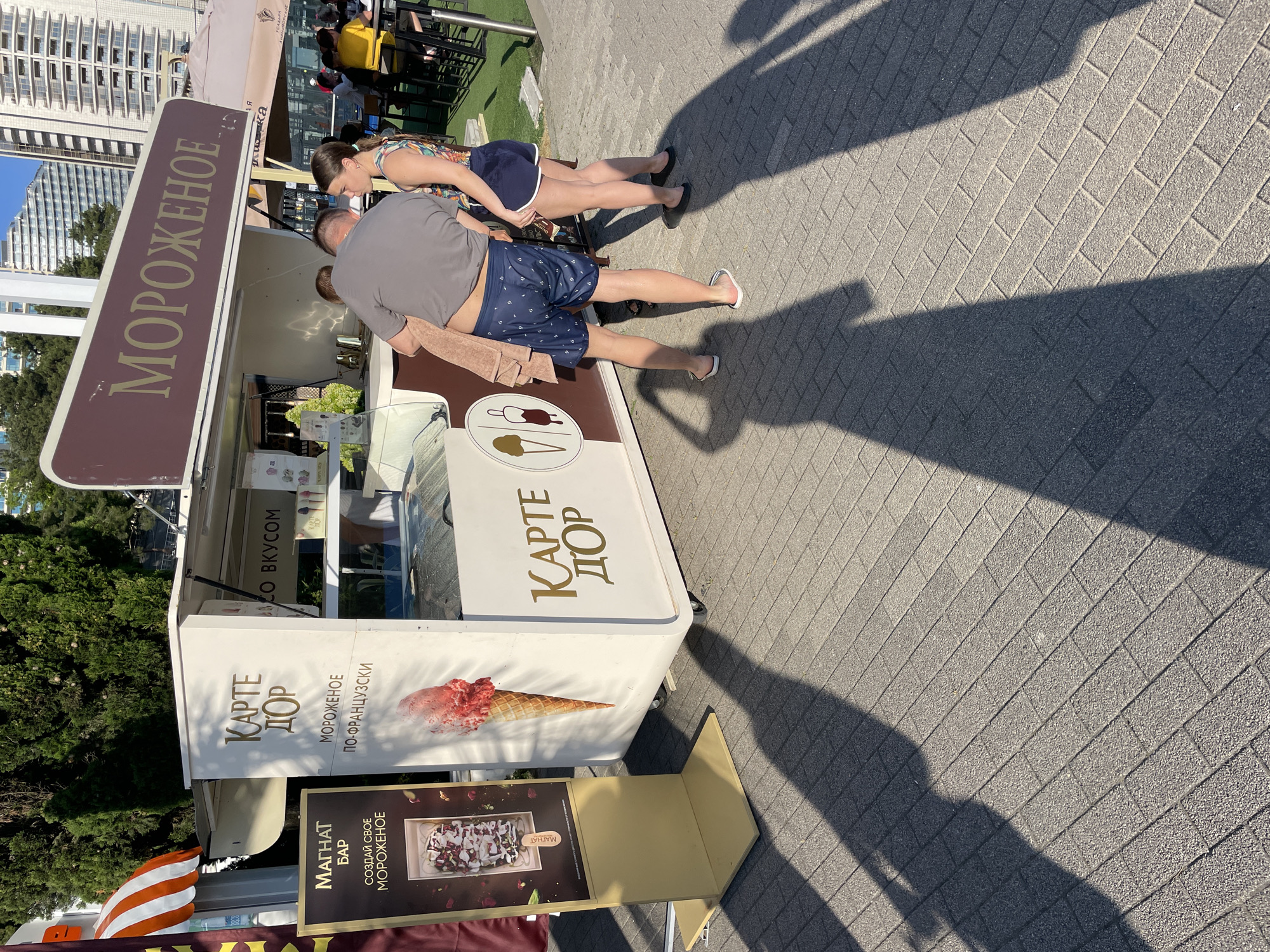 An outdoor ice cream kiosk is depicted in Sochi, Russia, under bright sunlight. The kiosk is white with a maroon awning and features the text "МОРОЖЕНОЕ" (ice cream) in large, gold lettering on the awning. Below this, the words "КАРТЕ ДОР" (Carte d'Or) are visible on the side of the kiosk, along with text indicating "МОРОЖЕНОЕ ПО-ФРАНЦУЗСКИ" (French-style ice cream). A large illustration of an ice cream cone adorns the side of the kiosk.
In front of the kiosk, three people are standing. A man, wearing a grey t-shirt and dark blue patterned shorts, is facing away from the camera and appears to be holding a beige towel. He is barefoot. To his right, a woman, wearing a patterned tank top and navy blue shorts, is looking towards him. To the far right, a young girl, also wearing a patterned tank top and navy blue shorts, is standing. Both individuals are also barefoot or wearing very flat shoes.
To the left of the kiosk, a display stand with a dark background shows an image of an ice cream dessert and the text "МАГНАТ БАР" and "СОЗДАЙ СВОЕ МОРОЖЕНОЕ" (Create your own ice cream).
The ground is paved with grey rectangular bricks, and long shadows of the people and the kiosk are cast across it, indicating the time of day is likely afternoon. In the background, lush green trees and modern white buildings are visible, suggesting a developed urban environment. The overall scene conveys a casual, summery atmosphere.