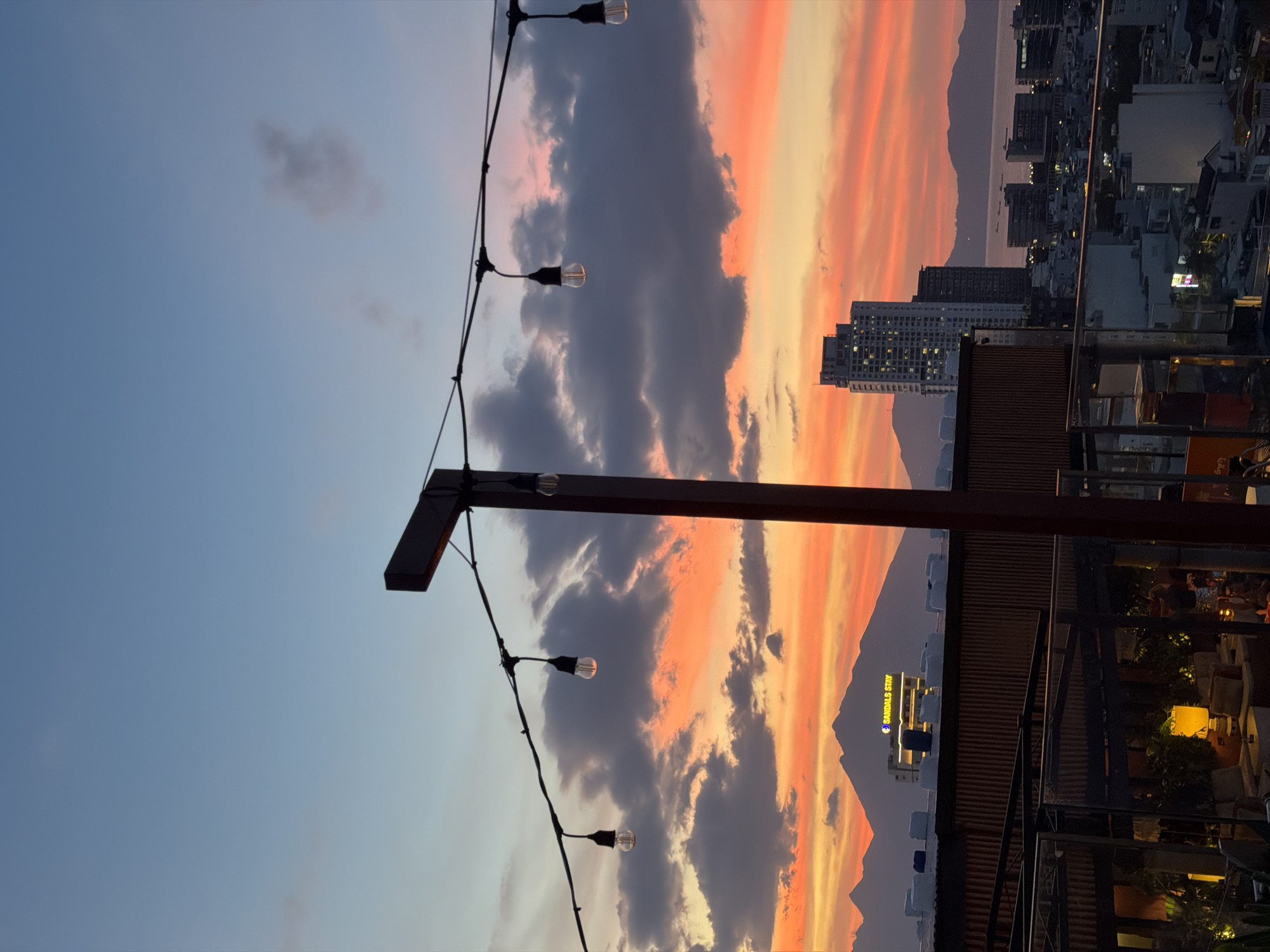 This is an outdoor, eye-level shot of a sunset over a city with mountains in the distance. In the foreground, a wooden beam and string lights with exposed bulbs hang over a rooftop restaurant or bar. The sky is filled with clouds illuminated by the sunset, displaying hues of orange, pink, and purple. The city skyline features several tall buildings, including a prominent skyscraper in the middle distance and a hotel with a lit sign that reads "SANDALS STAY" behind a mountain range. The water of the sea is visible in the background, with ships dotted across it. The overall atmosphere is serene and picturesque.