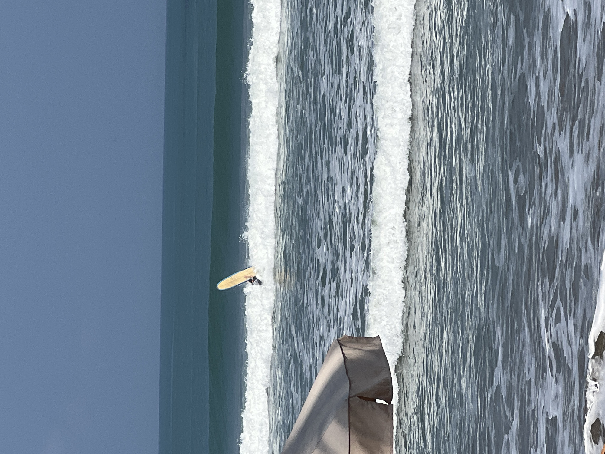 A surfer is attempting to ride a wave in the ocean off the coast of Kuta, Indonesia. The surfer is standing on a light-colored surfboard, which is angled vertically as the wave breaks. The surfer is wearing dark clothing. In the foreground, a portion of a large, taupe-colored beach umbrella is visible on the left side of the frame. The ocean is a deep blue-green, with several rows of white-capped waves. The sky is a muted, overcast gray.