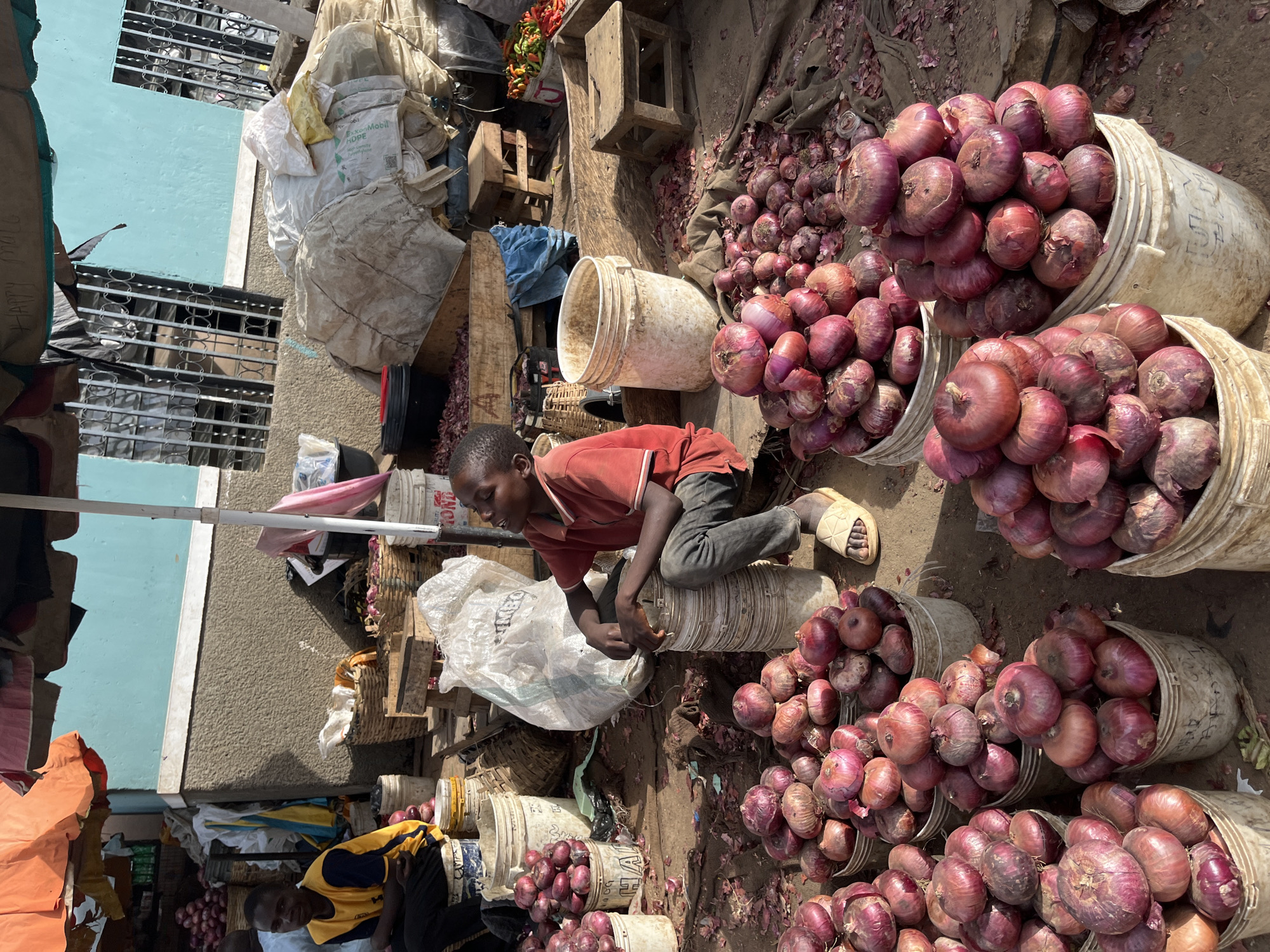 The image depicts a street market scene in Rafin Par, Nigeria, characterized by abundant displays of red onions. Two young individuals are present; one, a young boy in a red t-shirt and dark pants, sits on a white plastic bucket, looking upwards and to the right. The other, a young man in a yellow and blue shirt and black pants, is seated to the left, facing left. The ground is littered with onion skins. Various containers, including white plastic buckets and woven baskets, are filled with onions. Large white sacks and other goods are piled up in the background, alongside wooden stools and what appear to be market stalls with orange and pink coverings. A light blue wall forms the backdrop, with barred windows visible. The overall environment suggests an active marketplace under daylight.