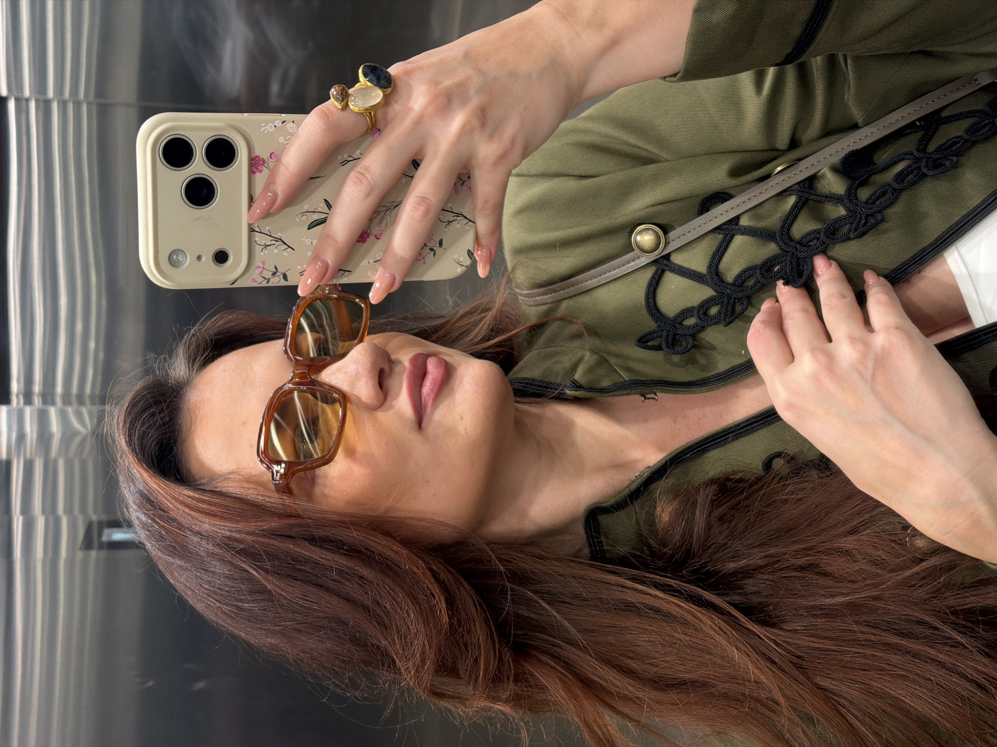 A woman is taking a selfie in what appears to be an elevator. She is wearing brown-tinted sunglasses, an olive-green jacket with black rope detailing and tan leather accents, and a large ring with a cluster of stones on her left hand. Her nails are long and manicured with a light pink polish. She holds a phone with a beige case decorated with a small floral pattern. Her long brown hair is styled in loose waves. The elevator has stainless steel walls with vertical lines, and based on the style of the jacket and accessories, the photo appears to have been taken in Belgrade, Serbia.